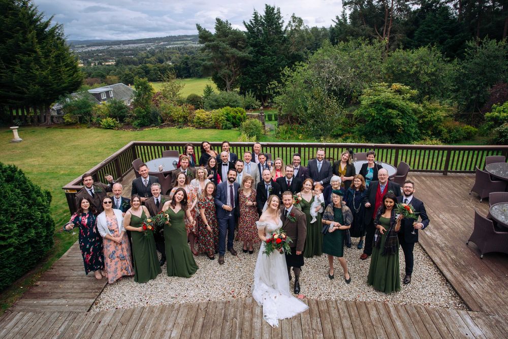 Wedding party gathered on a wooden deck overlooking scenic countryside landscape with rolling hills and trees.