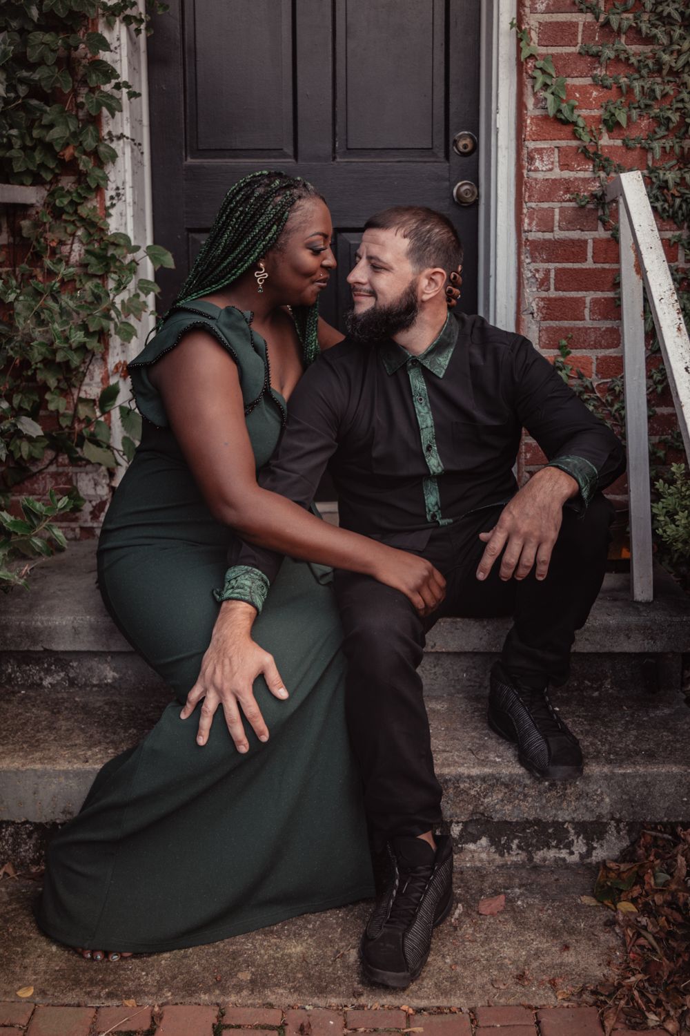 A romantic couple sits together on brick steps, with ivy growing on the wall behind them in moody lighting.