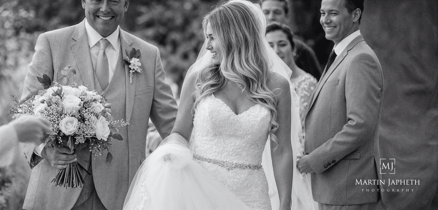 A candid black and white wedding moment shows the bride in a strapless gown laughing with groomsmen in light suits.