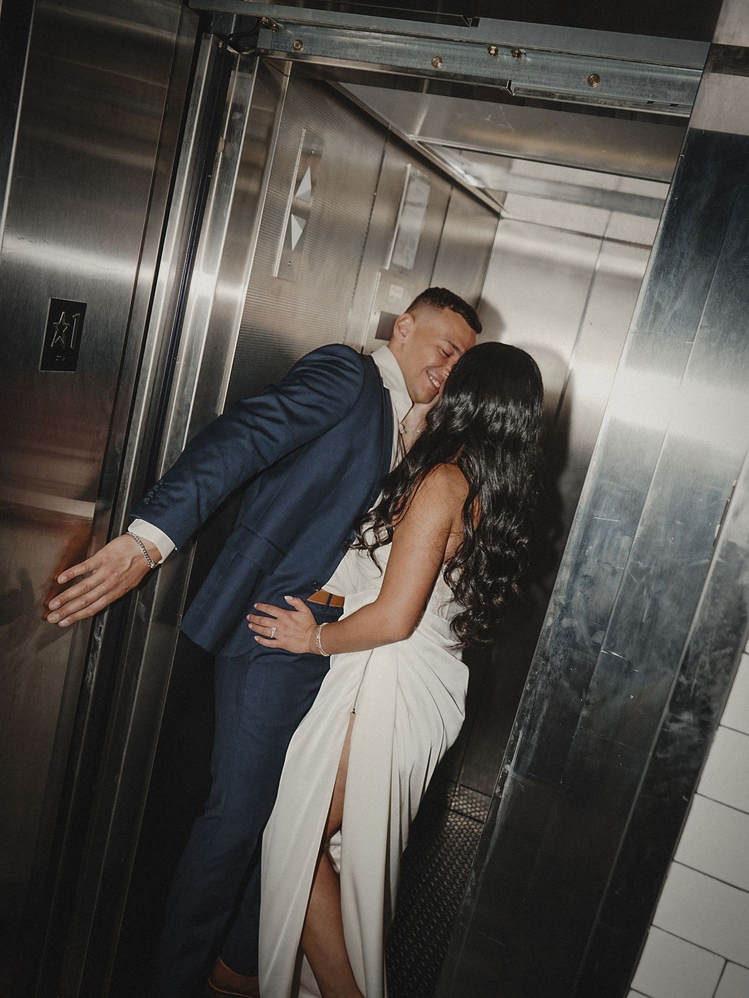Romantic couple sharing intimate moment in modern elevator wearing formal navy suit and white dress.