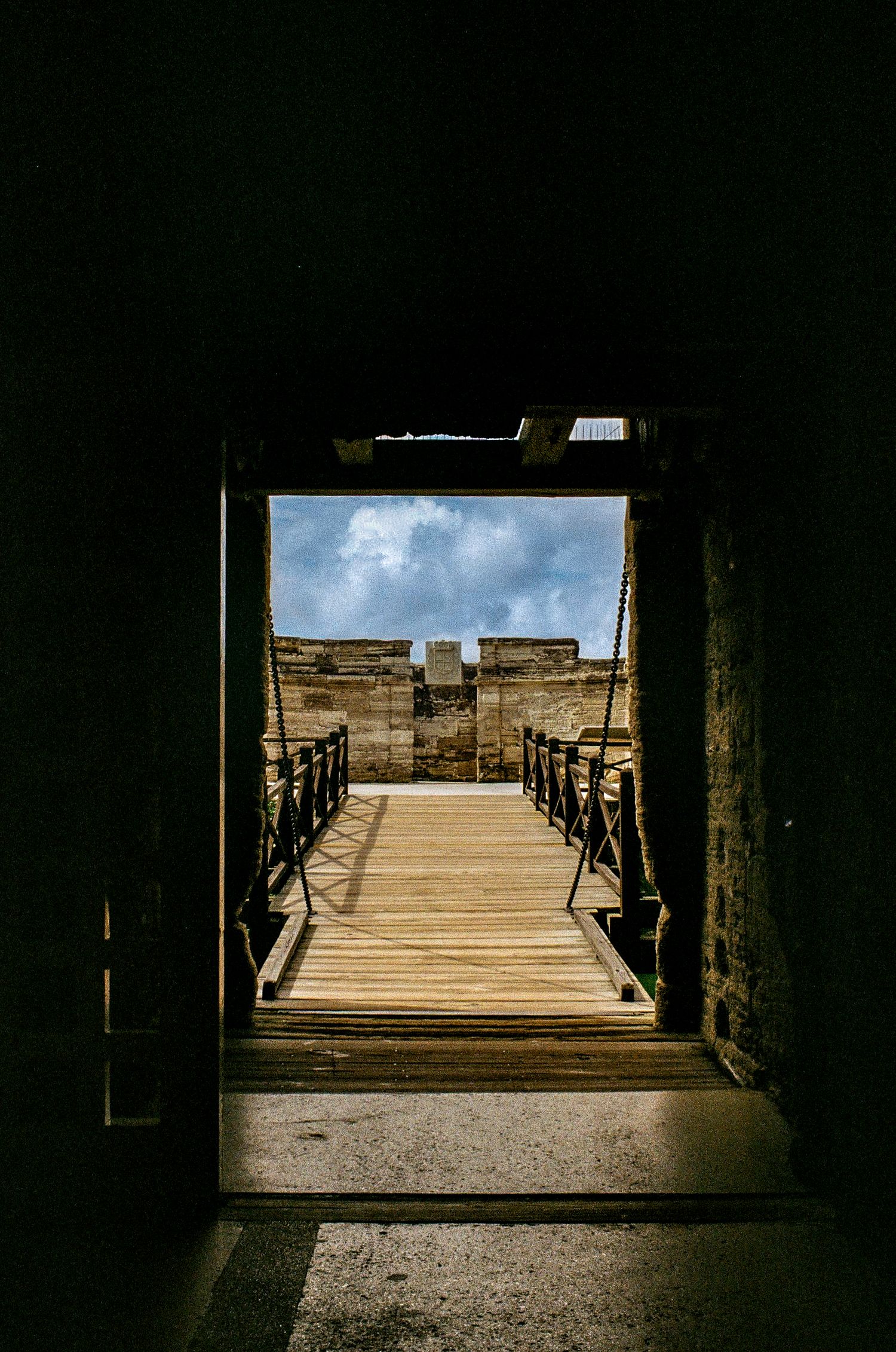 Wooden drawbridge entrance to ancient stone fortress walls under cloudy sky viewed through dark archway.
