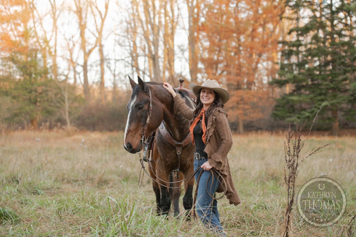 A rider in a brown coat poses with their horse in a fall meadow with trees in the background.