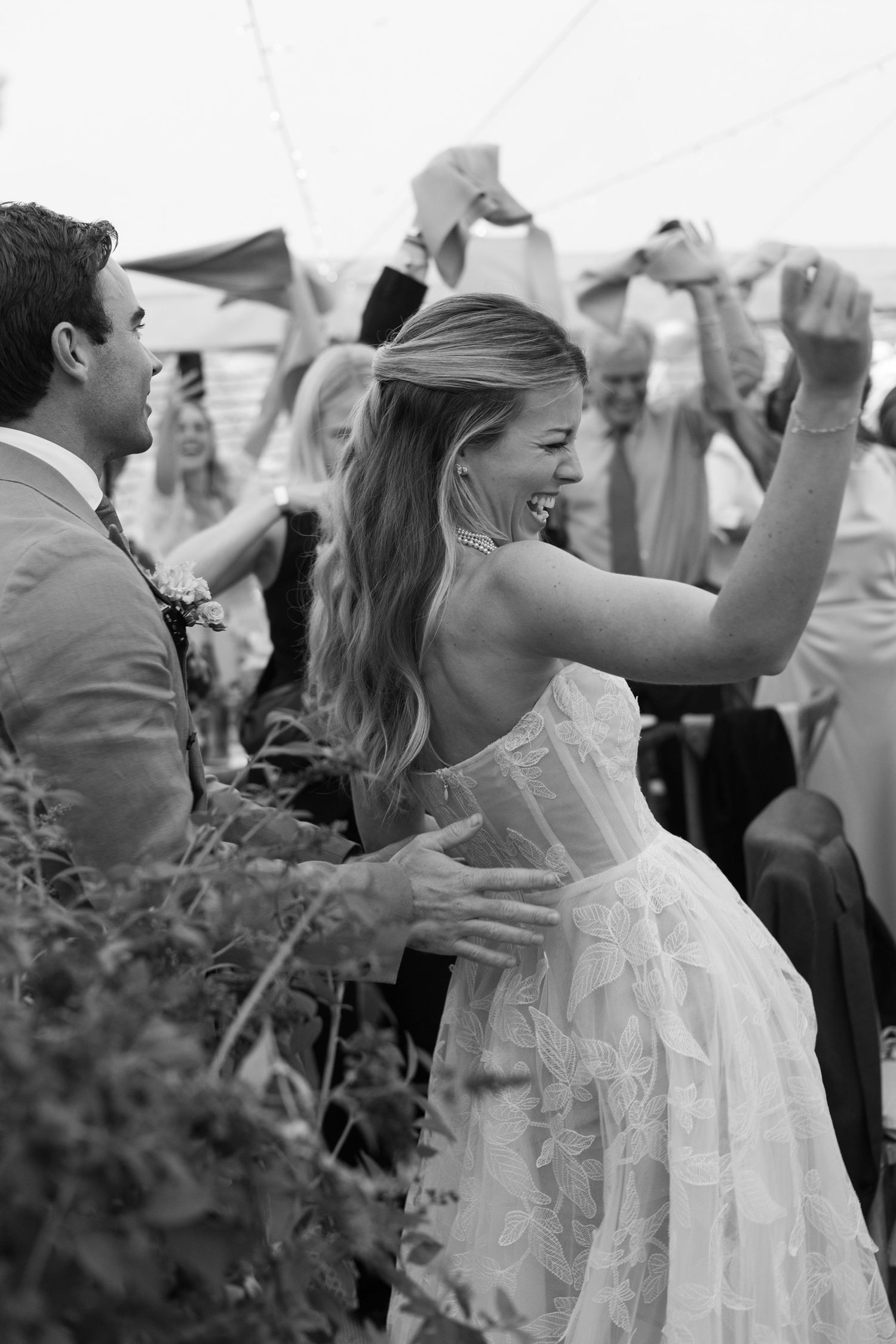 Black and white photo of joyful dancing at an outdoor wedding reception with a bride in a white lace dress.