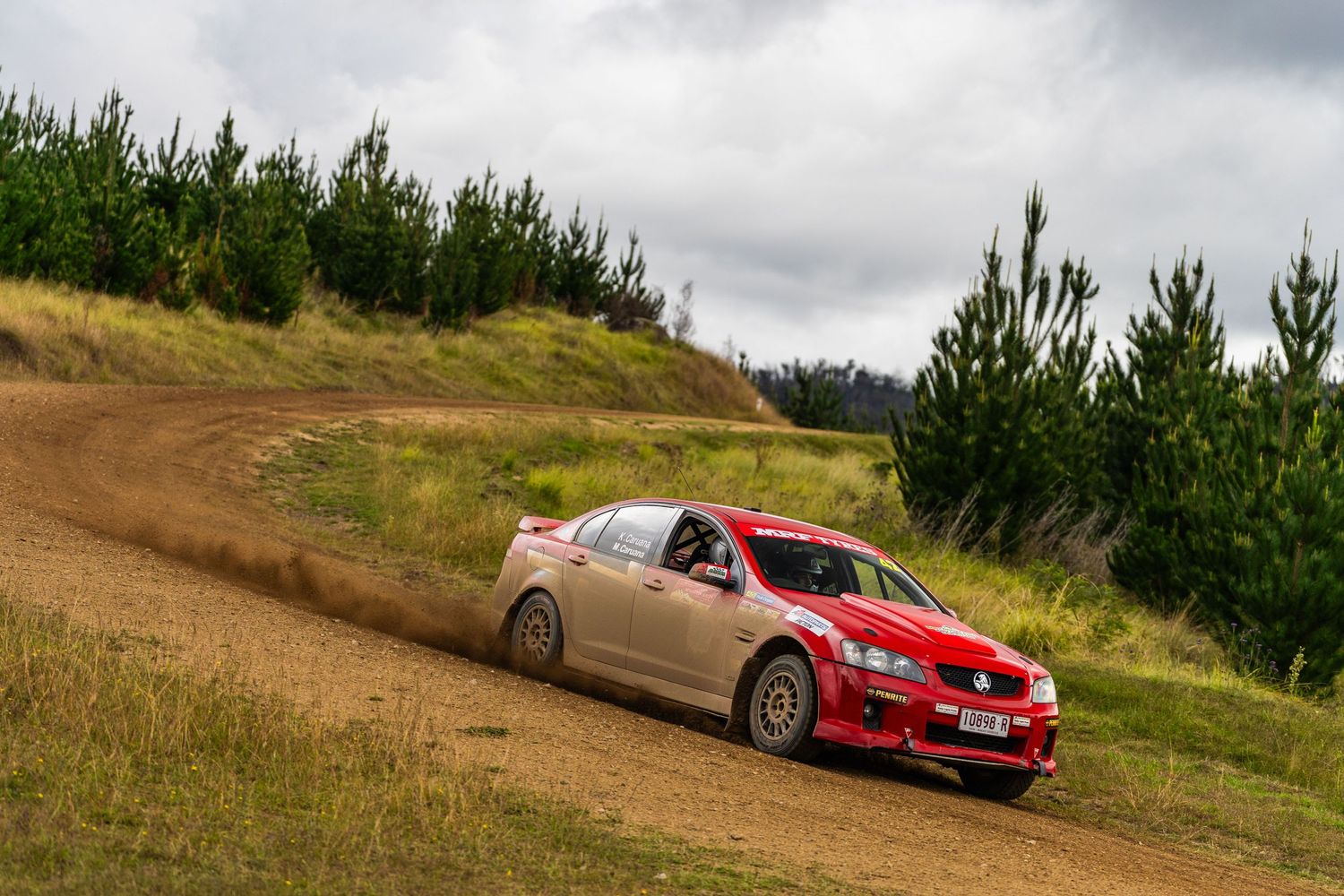 Red Subaru rally car races on dirt track through hilly pine forest terrain under cloudy sky.