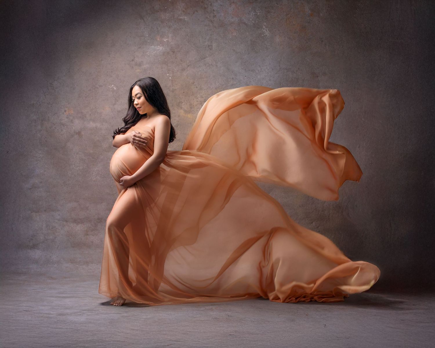 Maternity photograph of a woman standing looking at her belly while the gold silk chiffon she wears flys up against a beautiful backdrop.