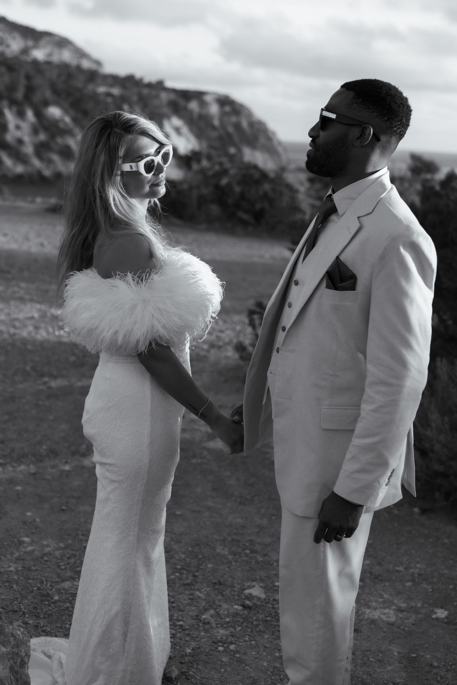 A black and white wedding photo of a couple outdoors, with the bride in a feathered dress and groom in a white suit.