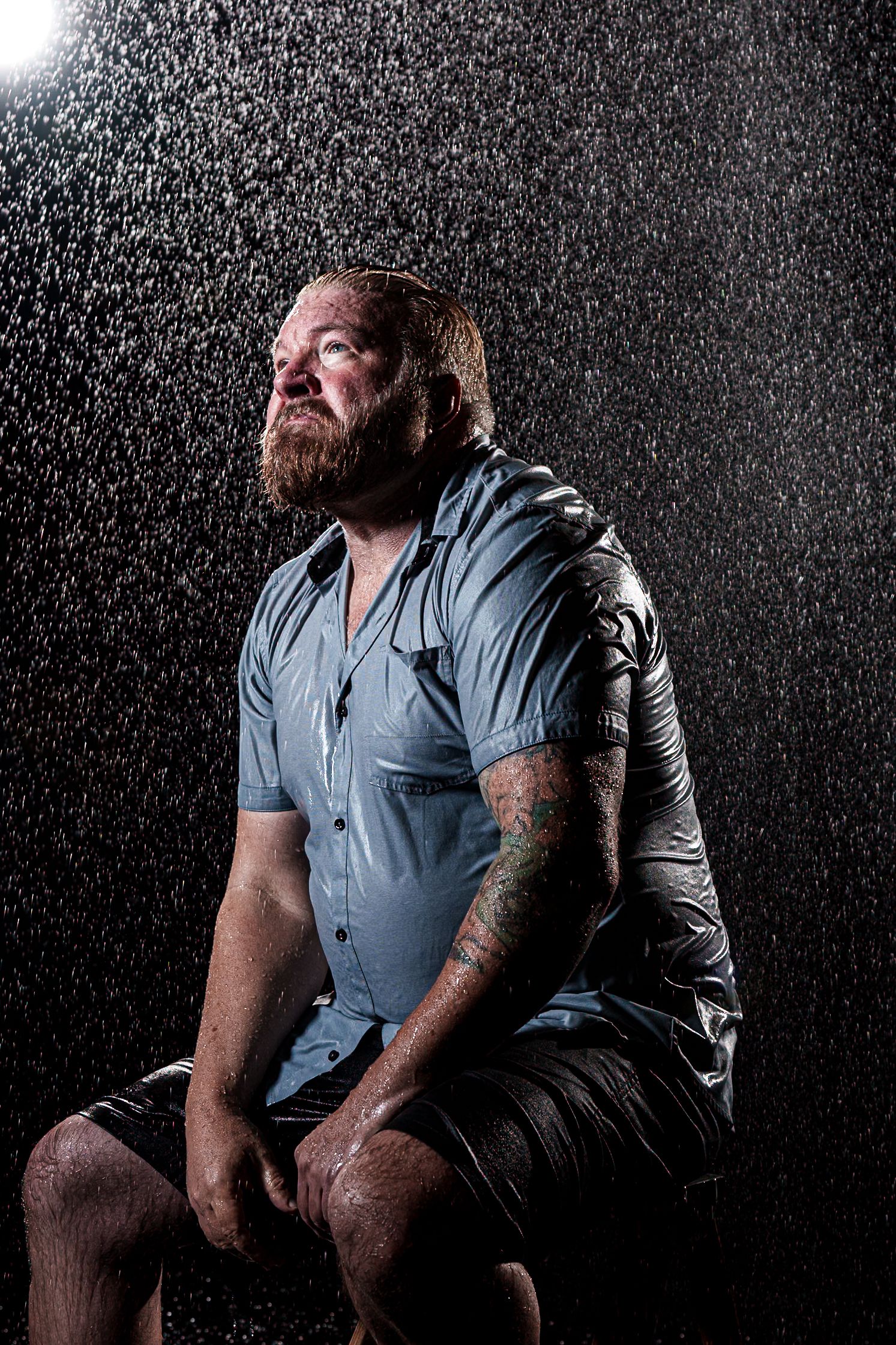 Dramatic portrait of a bearded person in a blue shirt sitting in water spray against dark background.
