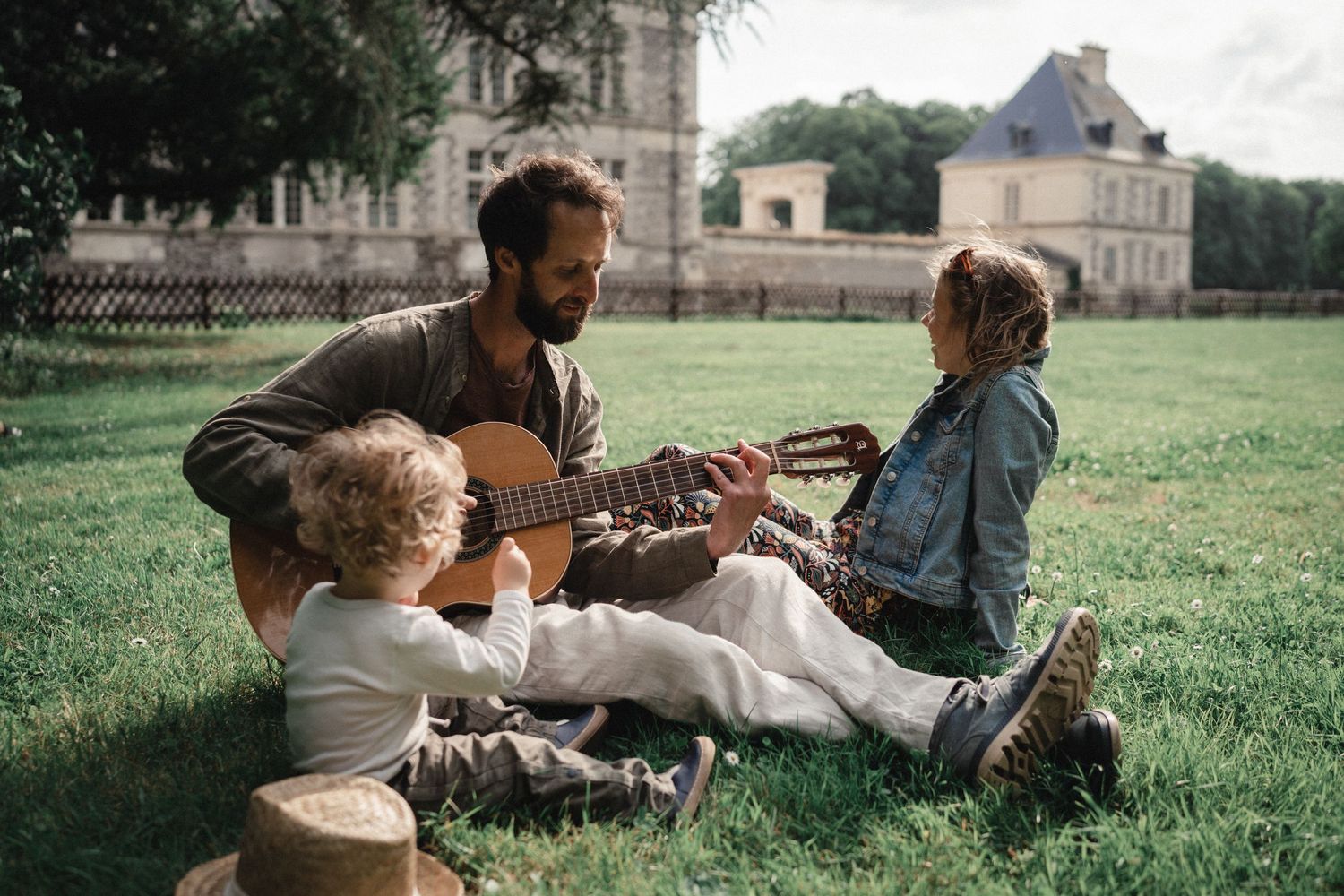 Famille profitant d’un moment musical en plein air tandis que quelqu’un joue de la guitare sur une pelouse, près d’un château.