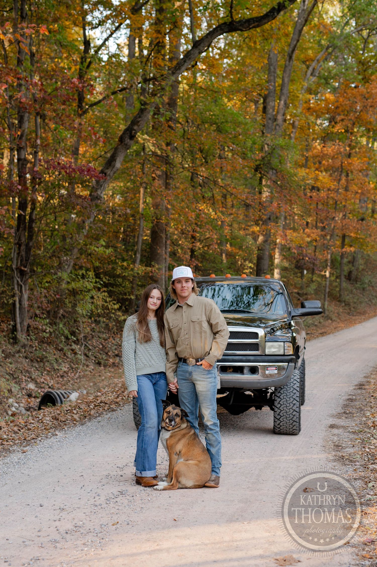 A brown dog joins companions posing in front of a lifted black Dodge Ram on an autumn forest trail.
