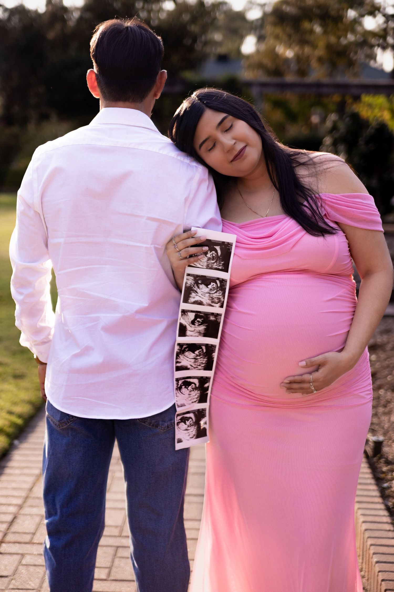 Expecting couple in white shirt and pink dress poses with ultrasound photos on brick pathway at Mercer Botanic Gardens in Houston Texas