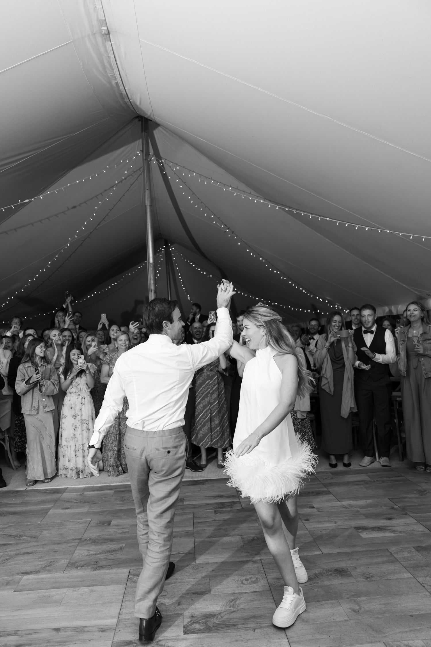 A couple dances energetically under a tent decorated with string lights while wedding guests watch from the sidelines.