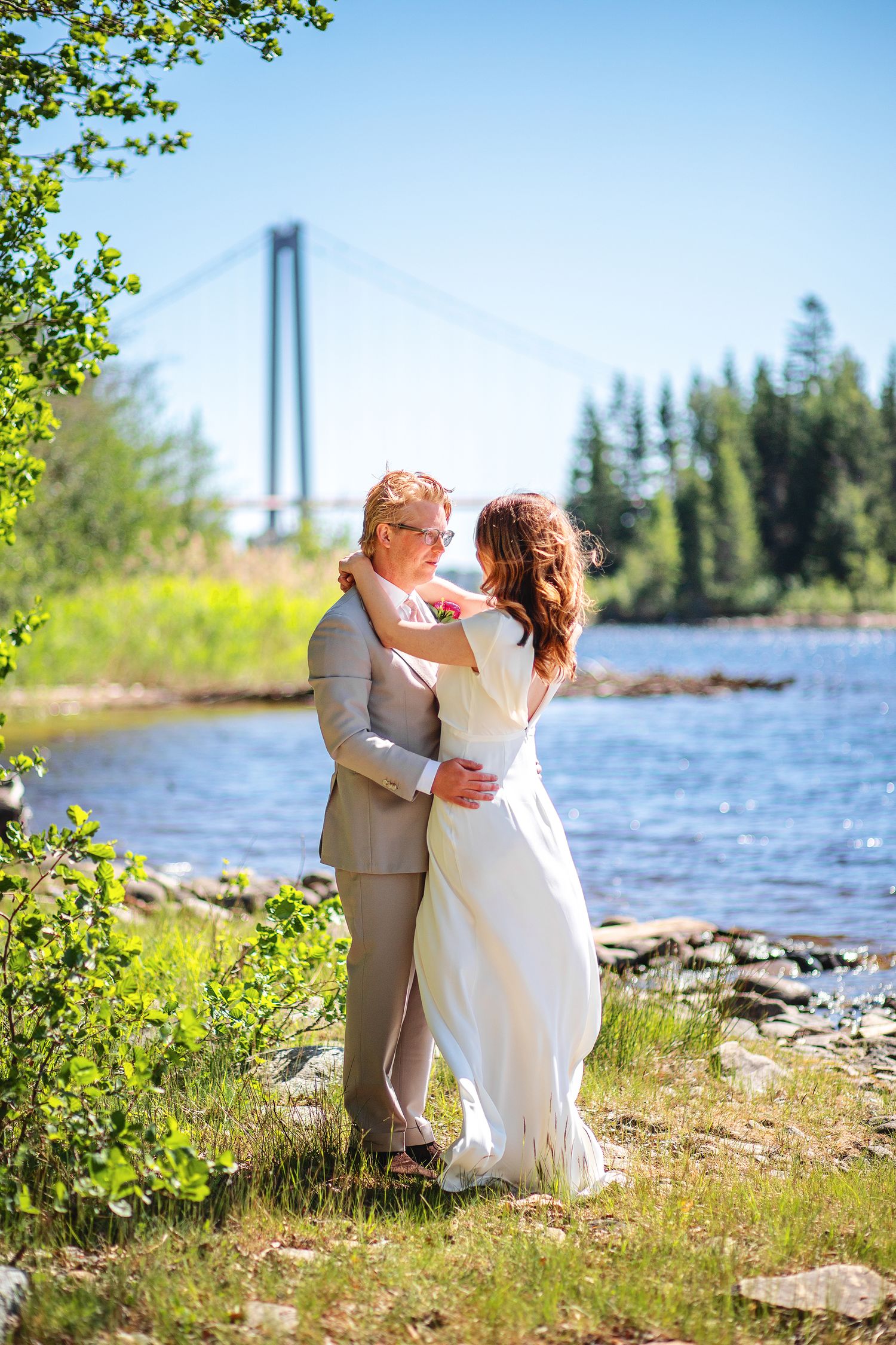 Wedding couple embracing by a riverside with a suspension bridge and green trees in the background on a sunny day.