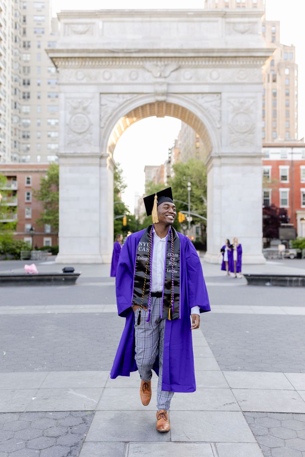 Graduate wearing purple graduation robe and decorated stole walks under historic archway.