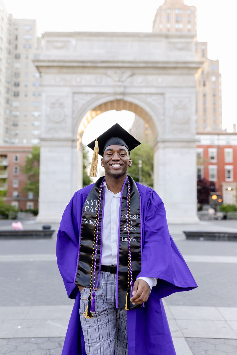 NYU graduate in purple regalia smiles proudly in front of Washington Square Arch.