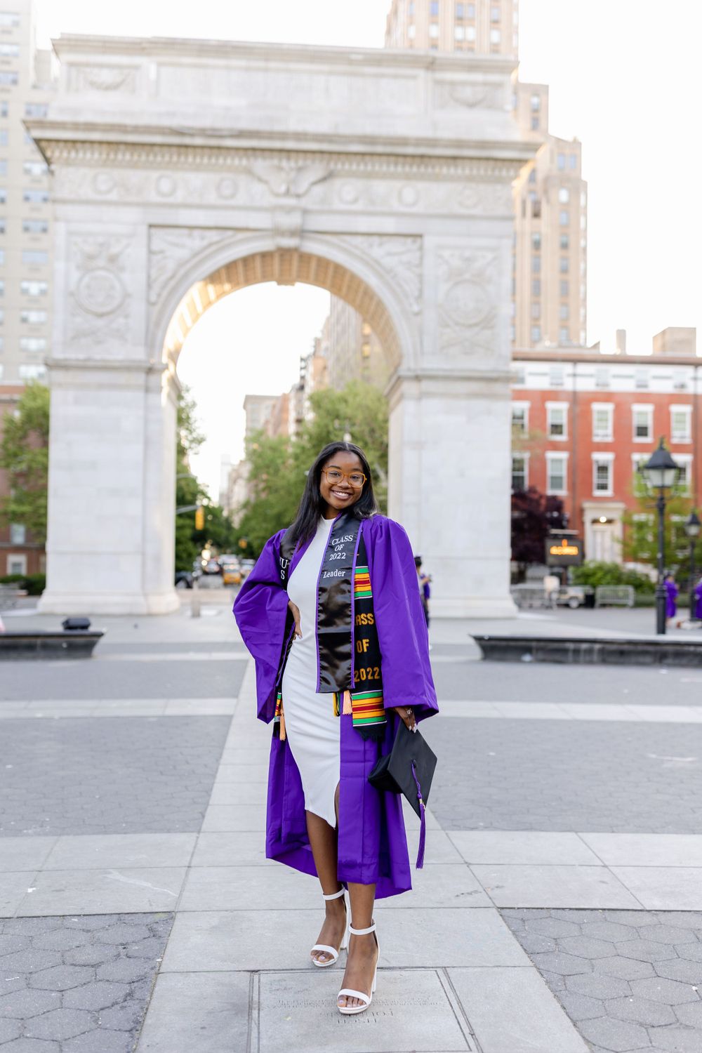 Graduate in purple gown and white dress poses elegantly at Washington Square Park.