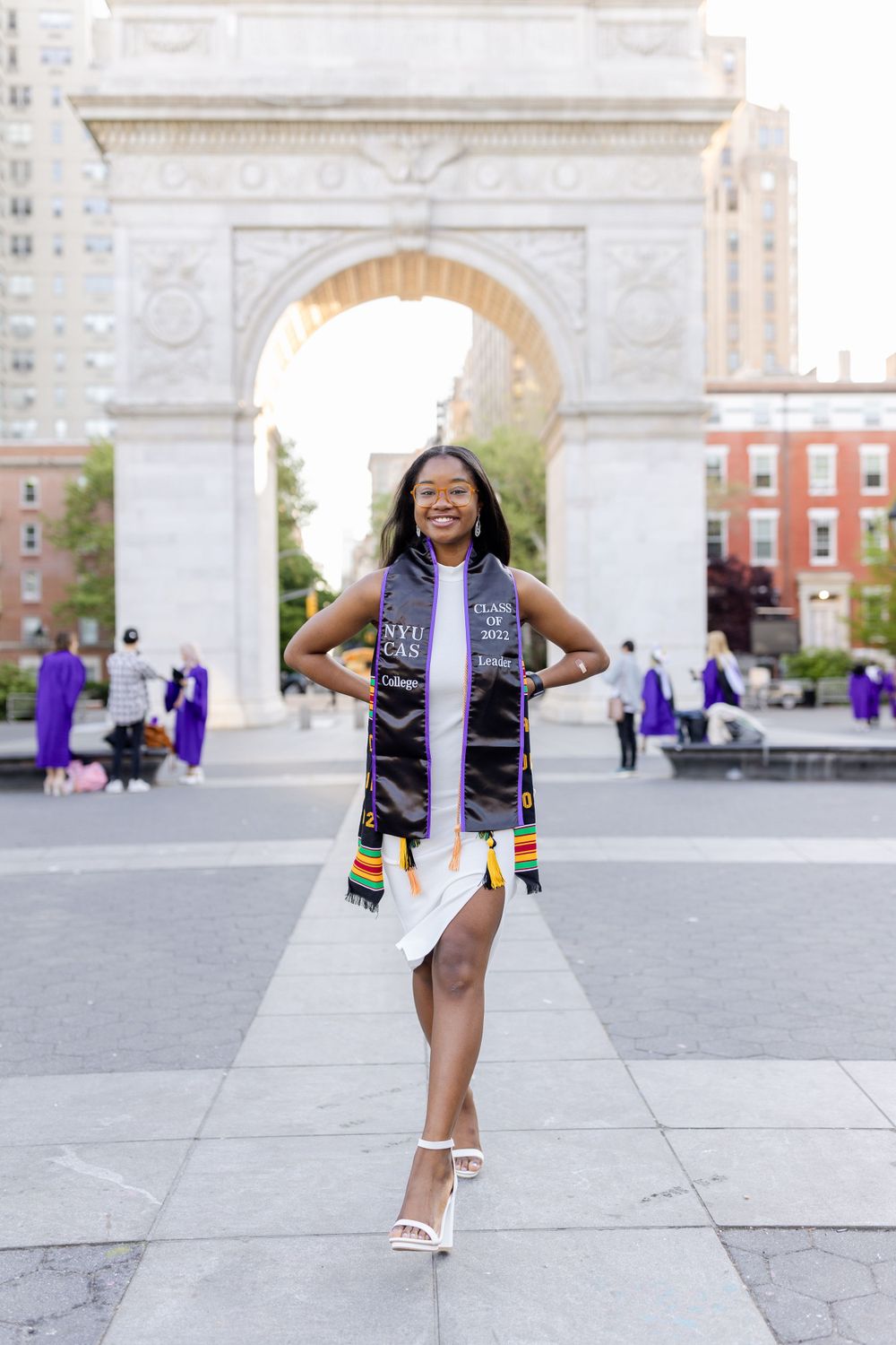 Graduate in white dress and purple stole poses confidently in front of historic arch.