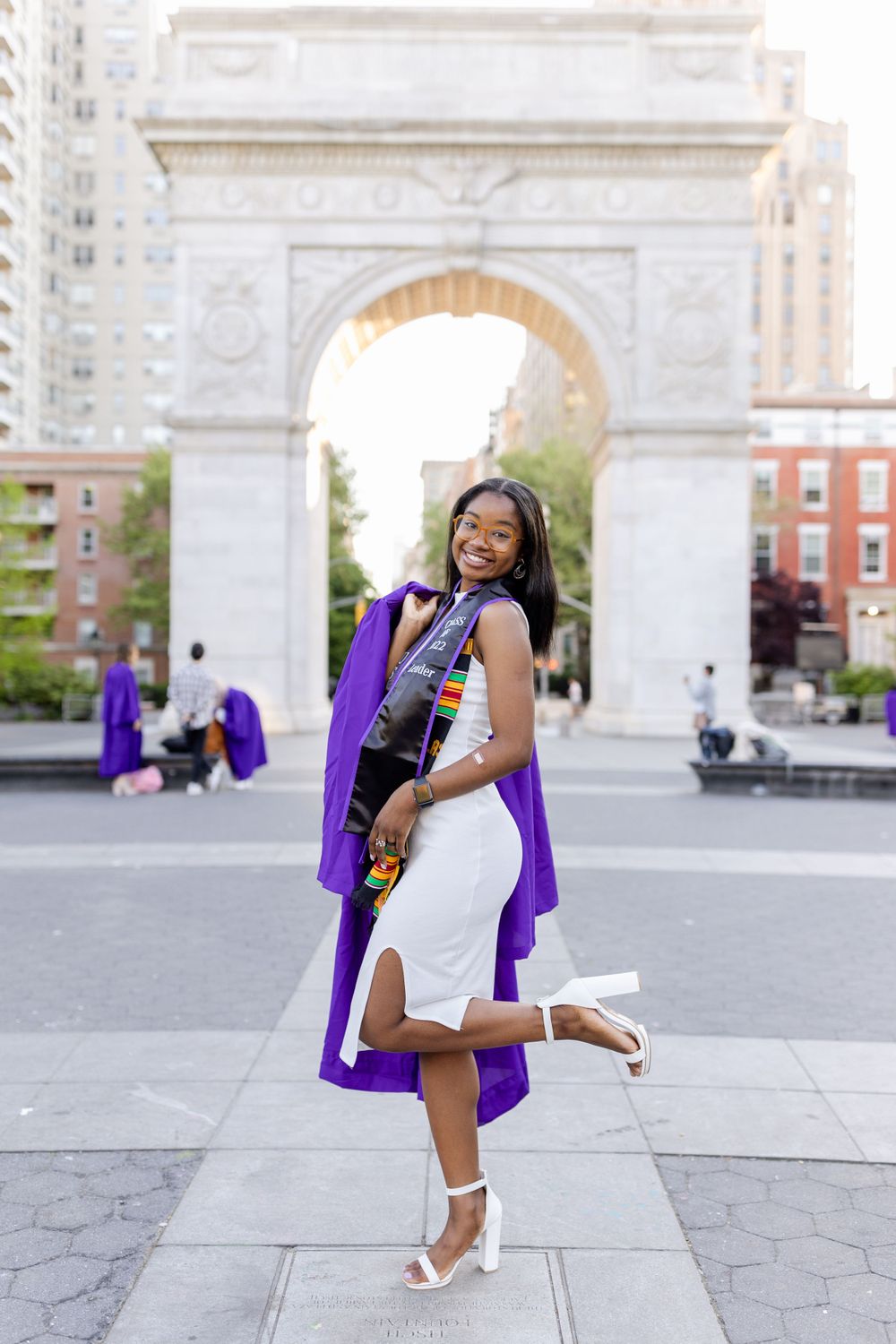 Graduate strikes playful pose in white dress and purple graduation gown at Washington Square Park.