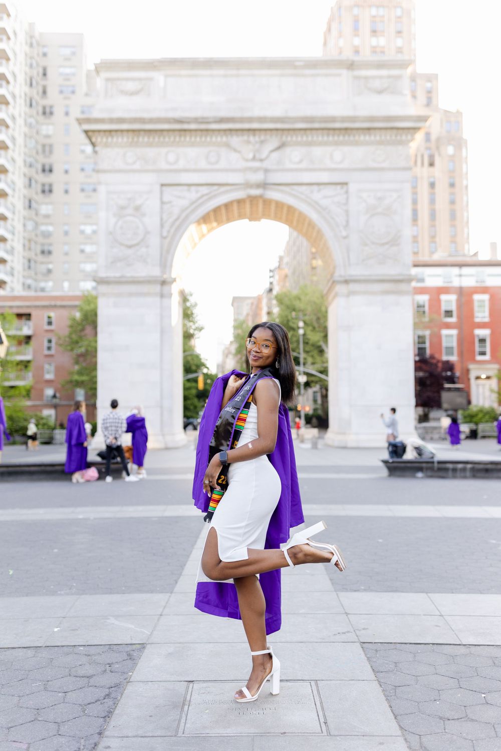 Graduate in white dress and purple graduation gown twirls joyfully at Washington Square Park.
