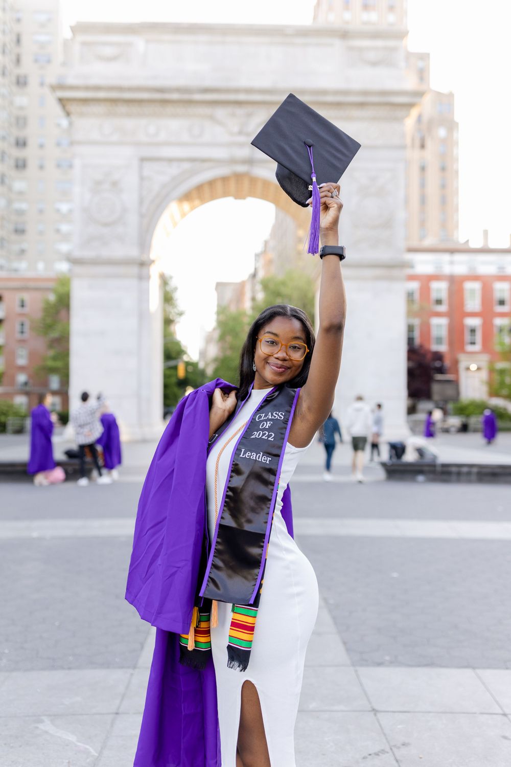 Graduate triumphantly raises graduation cap while posing at Washington Square Arch.