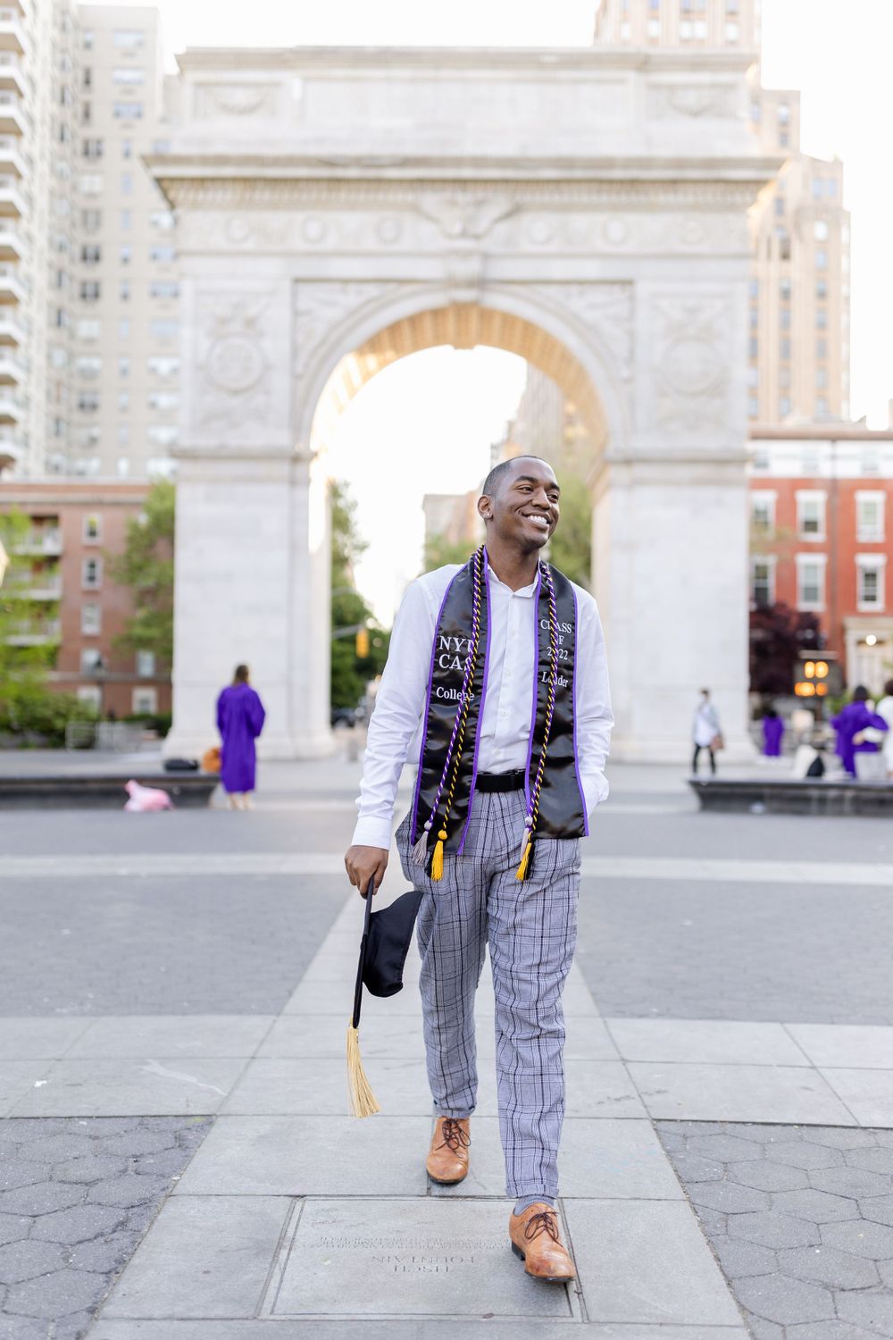 Graduate in plaid pants and graduation stole walks confidently at Washington Square Park.