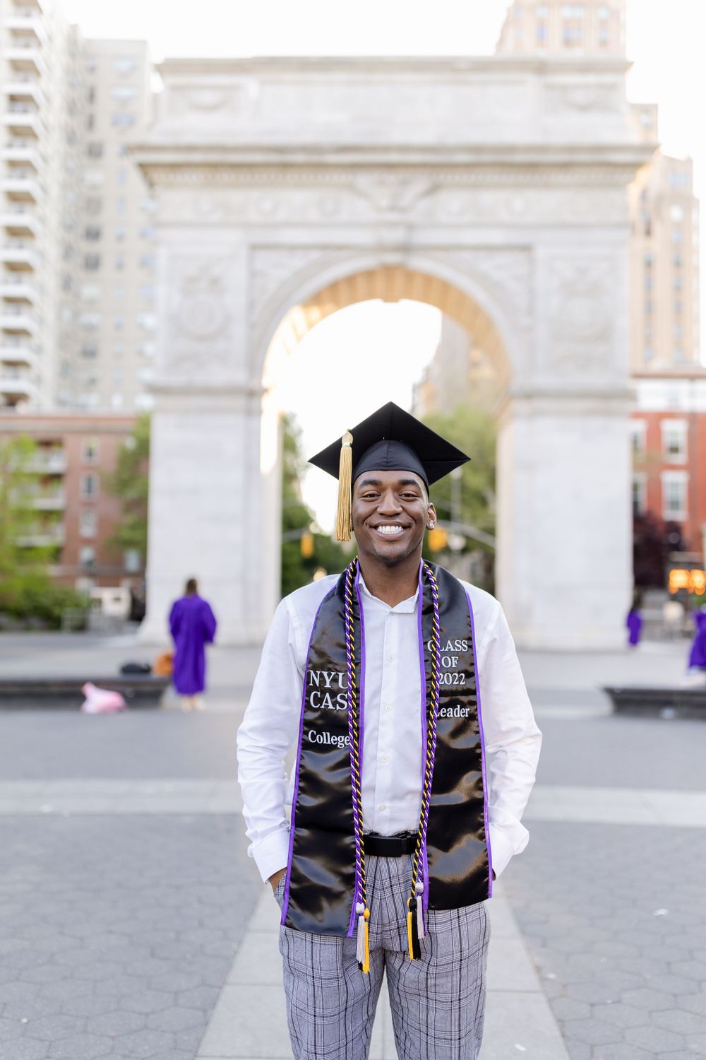 Graduate in plaid pants and graduation cap poses at Washington Square Park arch.