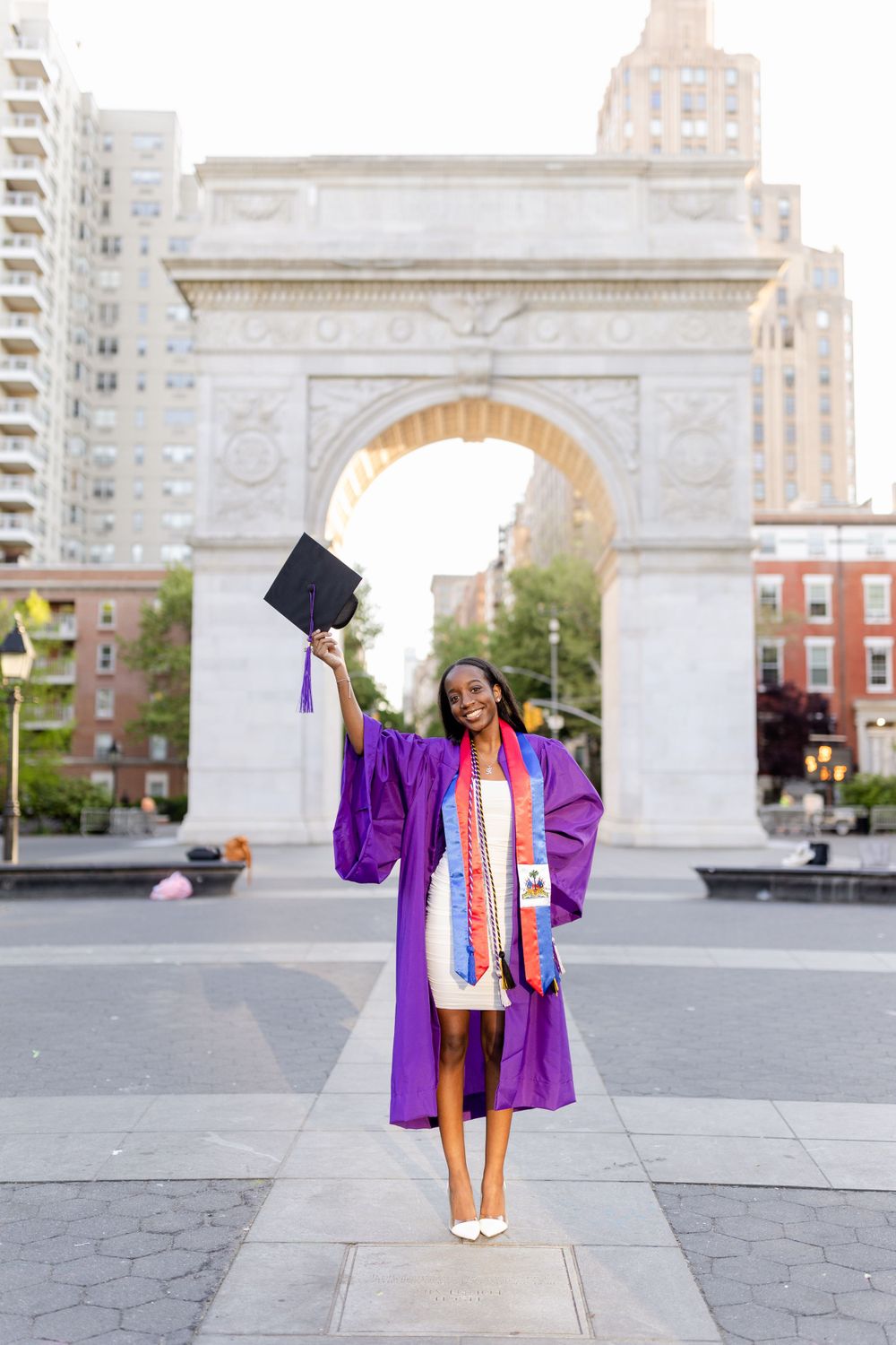 Graduate in purple cap and gown celebrates by jumping with diploma at Washington Square Arch in NYC.
