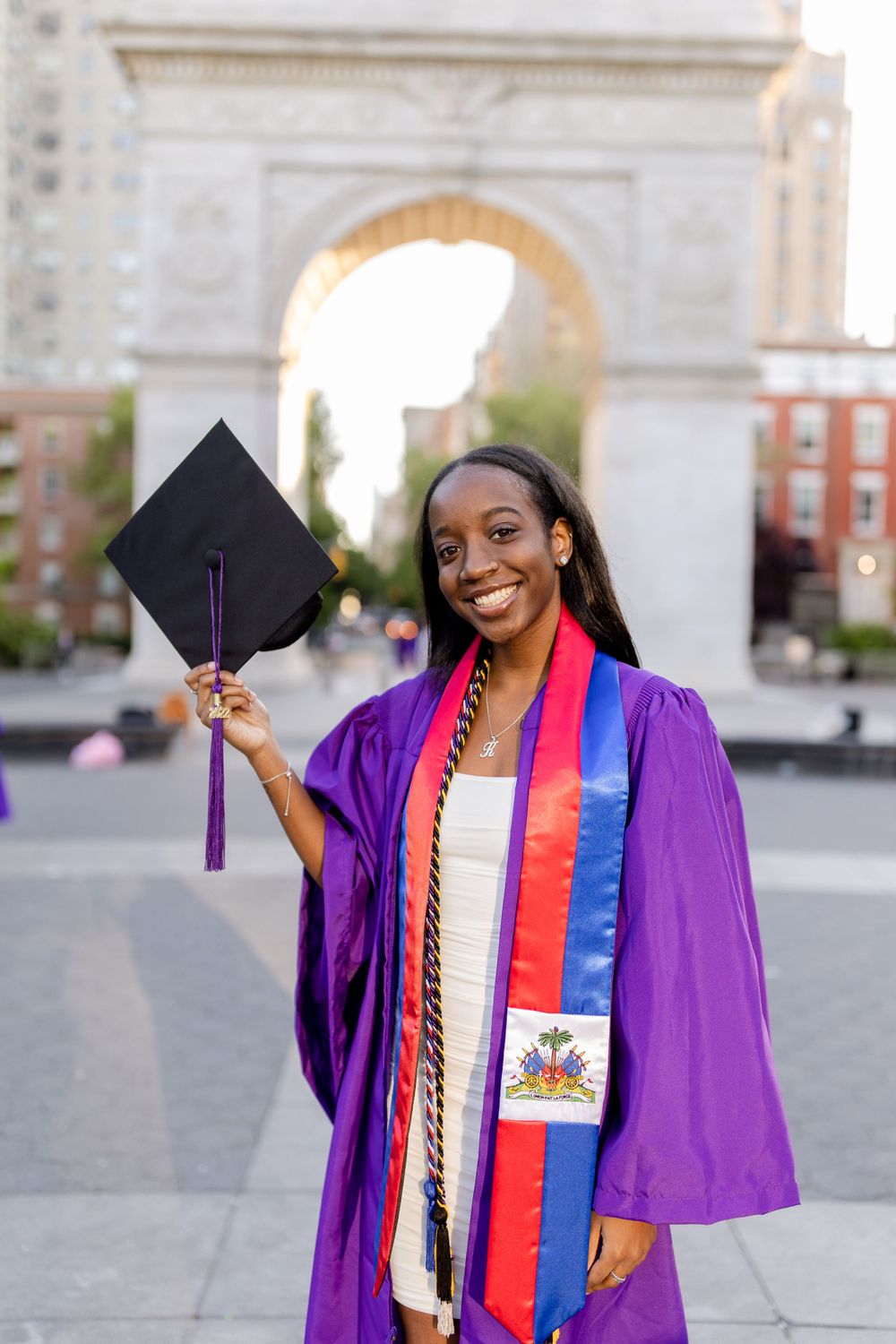 Graduate in purple robe with Haitian flag sash holds cap while smiling at Washington Square Park.
