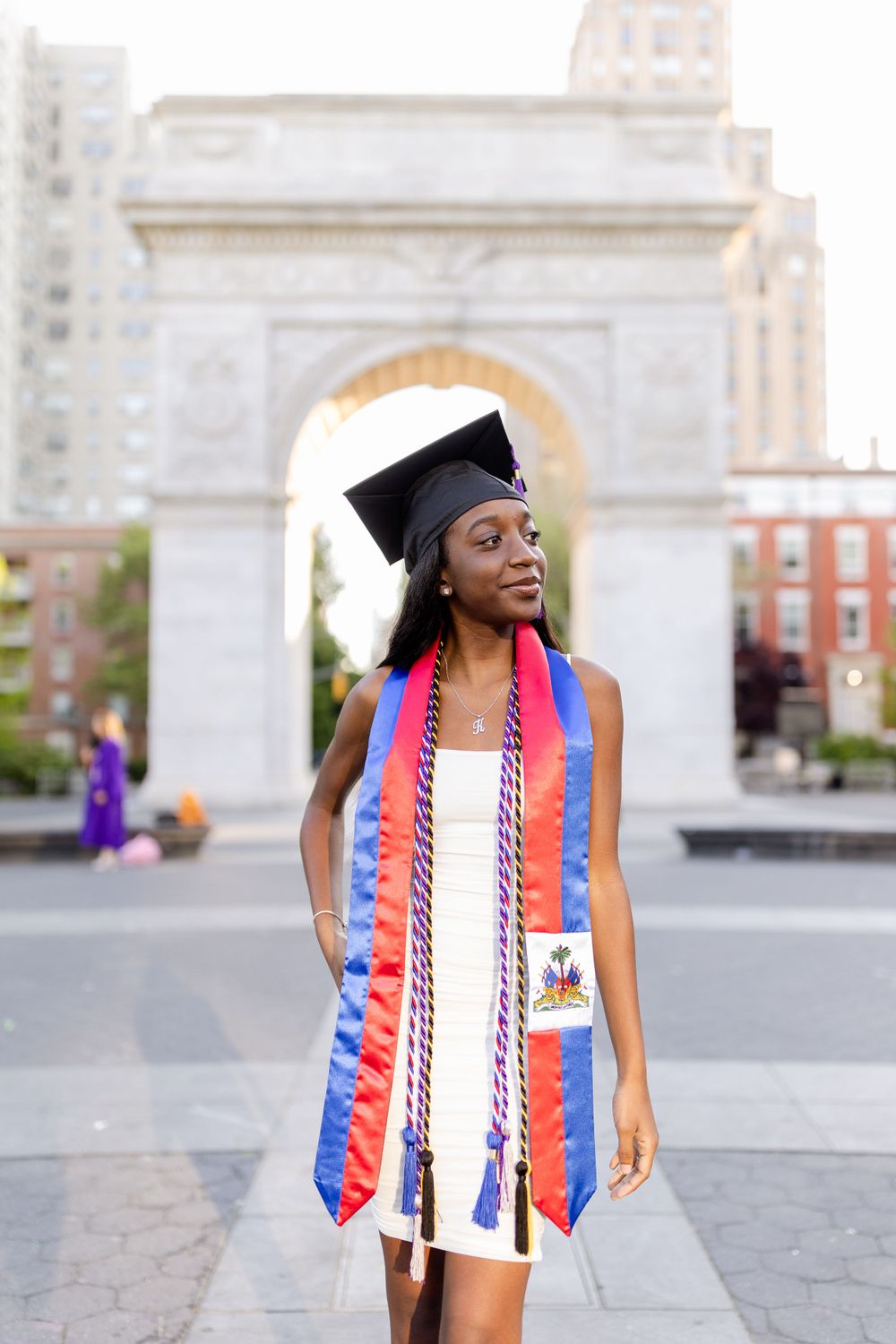 Graduate wearing decorative stole and cap poses in front of Washington Square Arch.