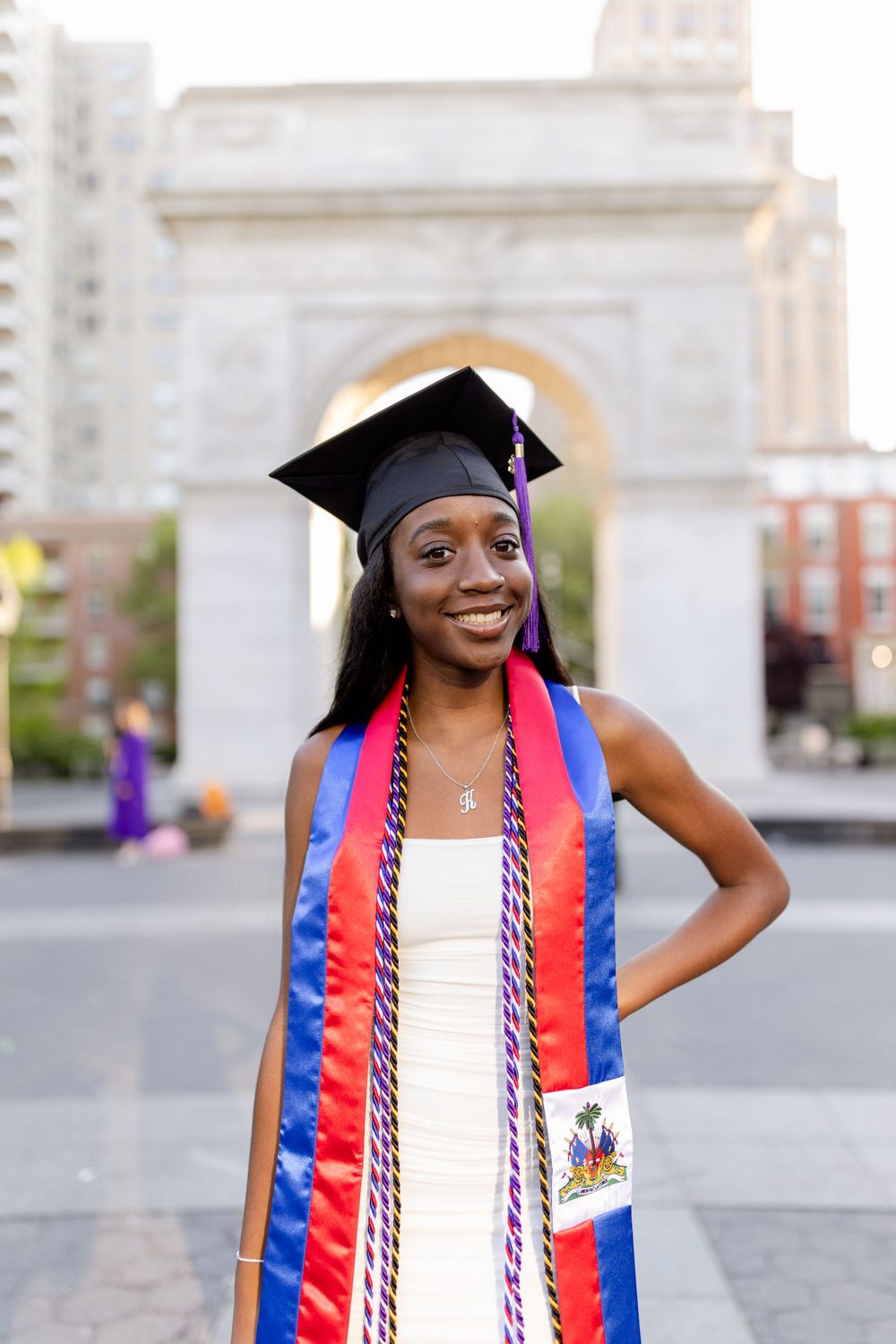 Graduate in academic regalia with special stole smiles in front of iconic NYC arch.