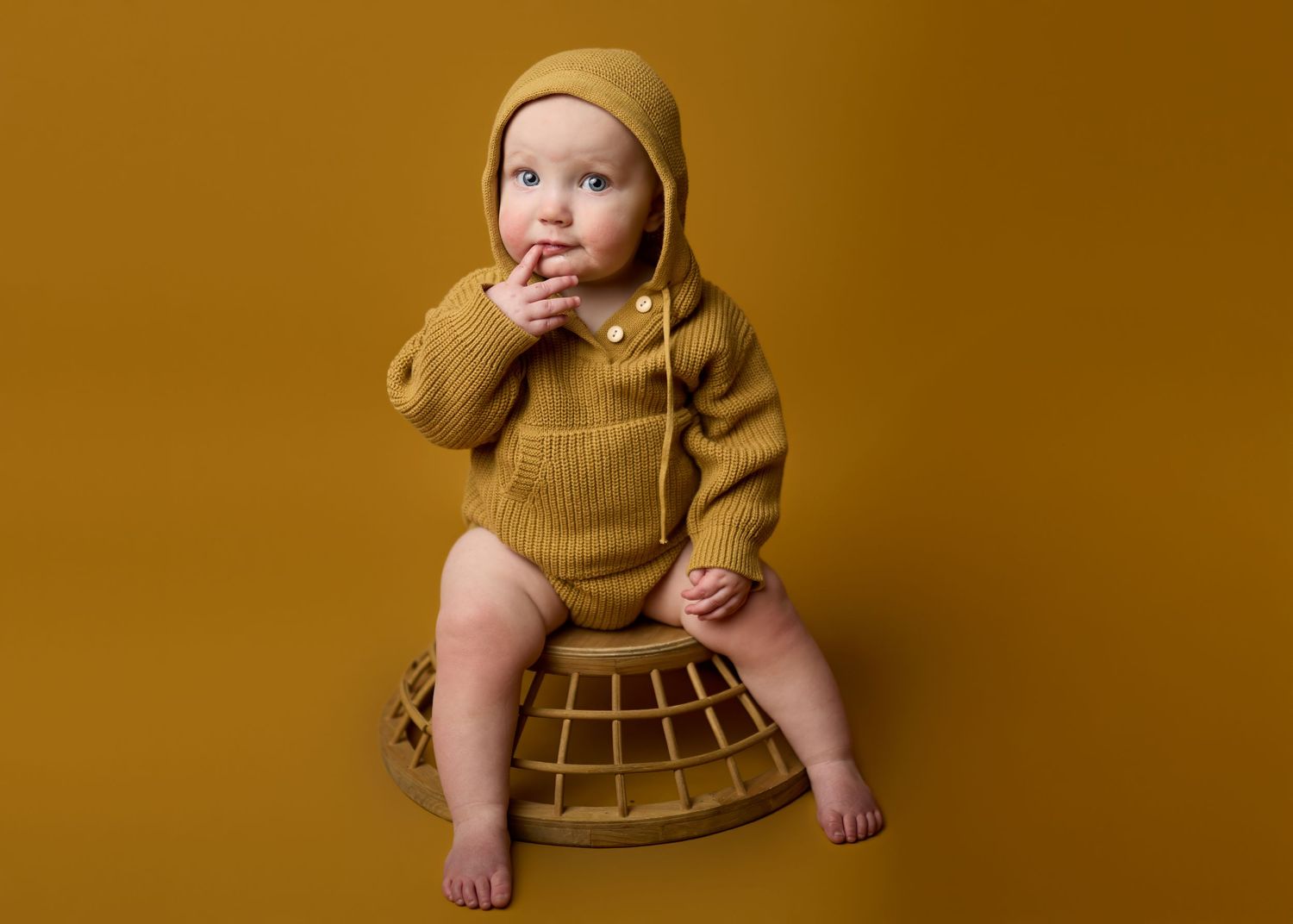 A baby in a mustard yellow sweater sits on a woven wicker stool against a warm brown background.