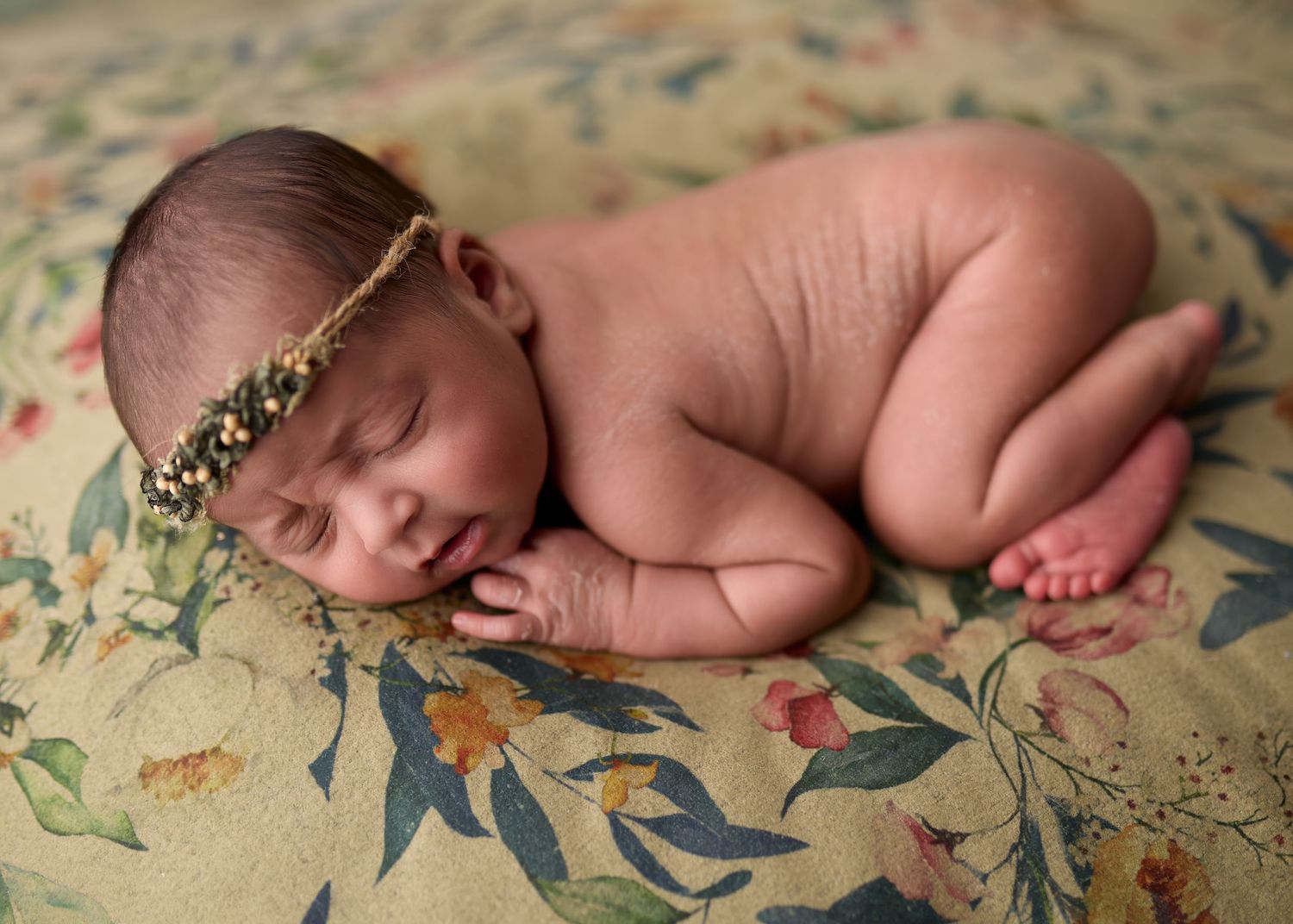 Peaceful sleeping newborn wearing delicate floral crown rests on floral patterned fabric.