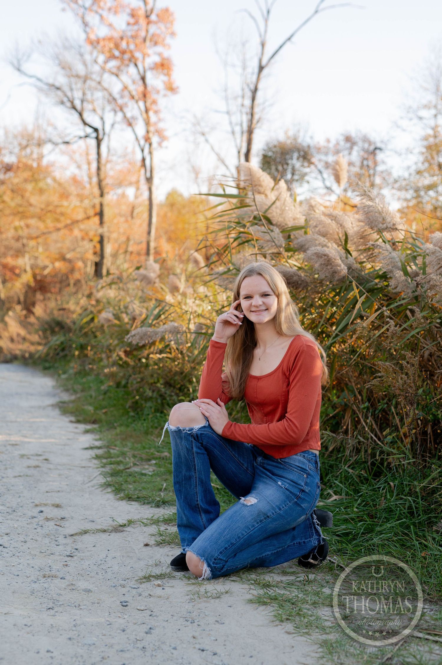 Person in red sweater kneeling on dirt path surrounded by autumn vegetation.