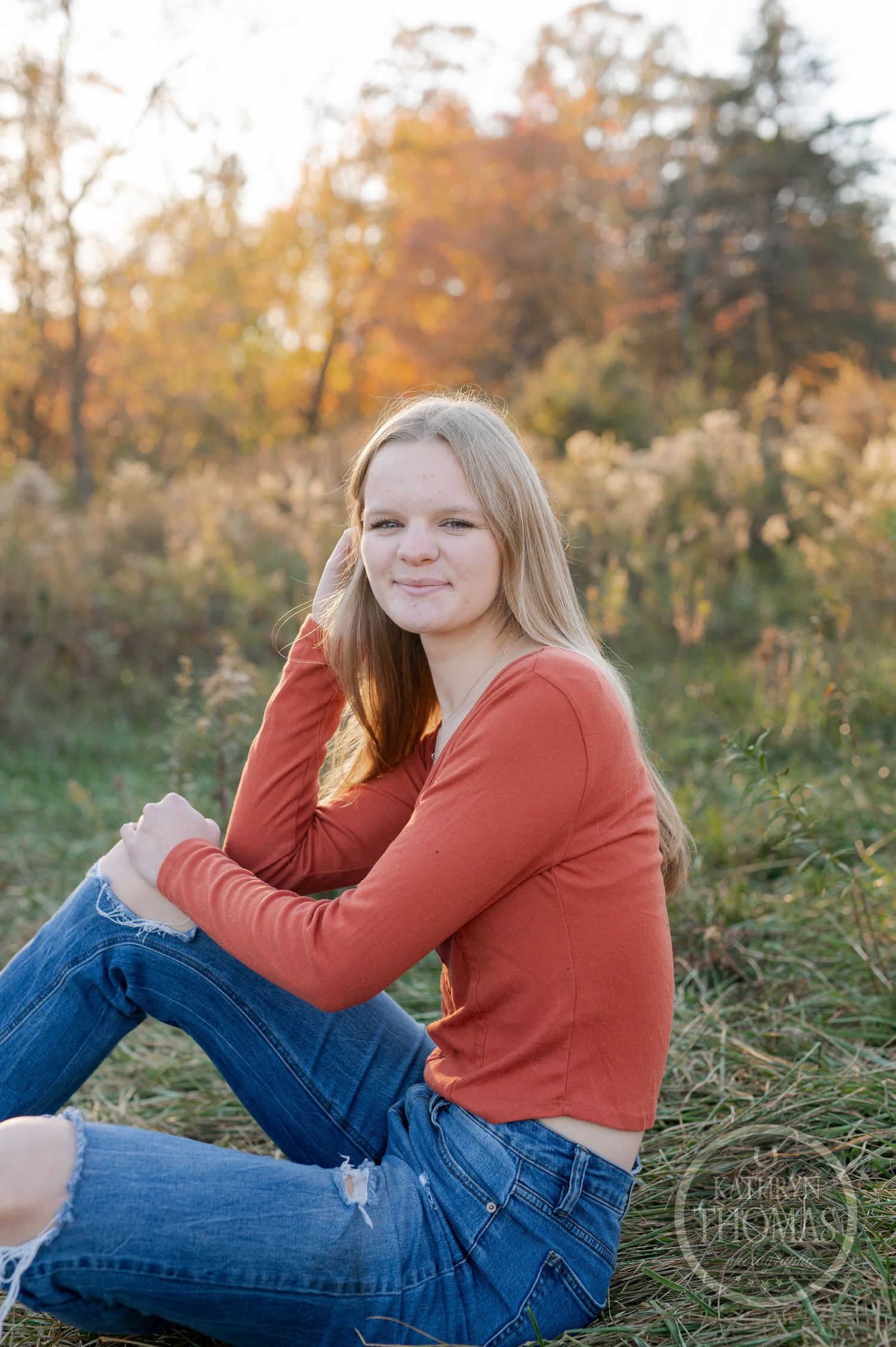 Relaxed outdoor photo featuring someone in autumn attire sitting on grass with fall foliage in background.