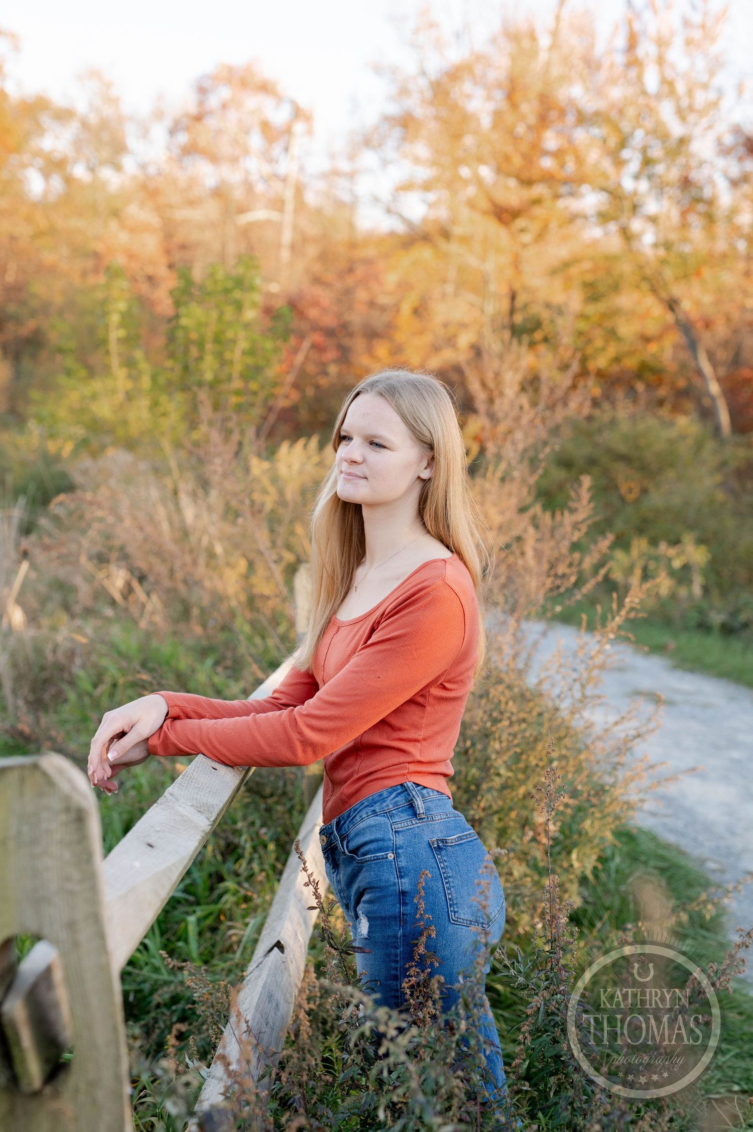 Someone poses casually against a wooden railing on a nature trail surrounded by autumn vegetation.