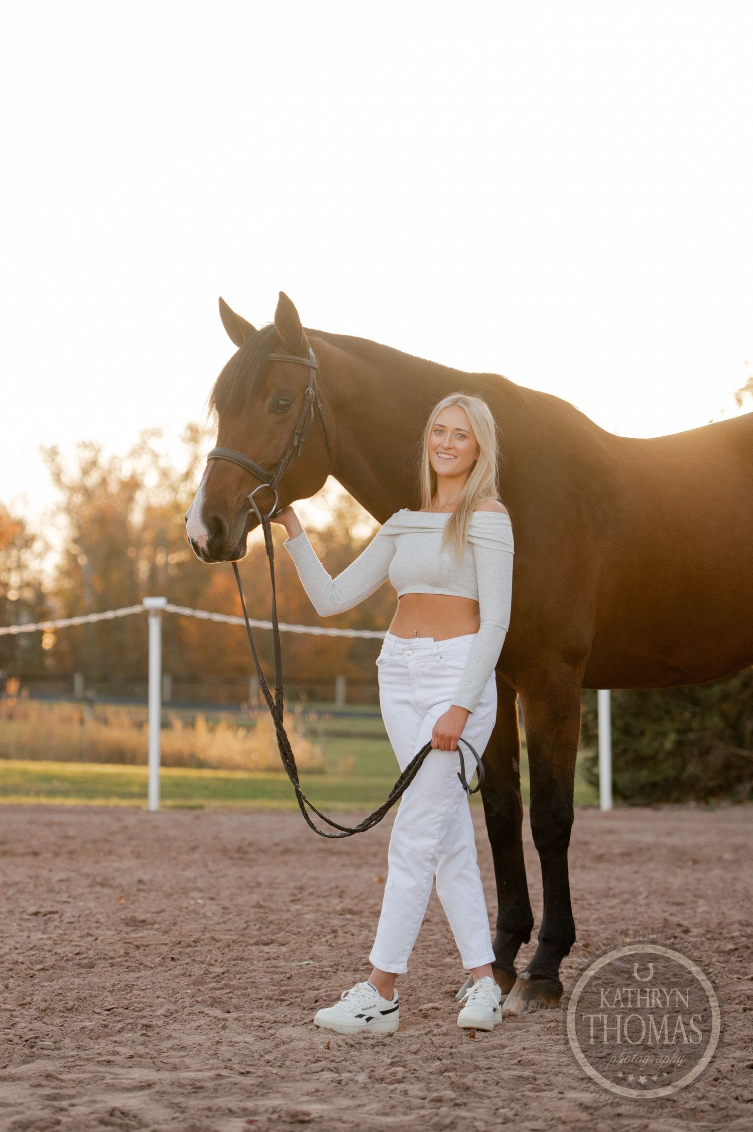 A person poses with a bay horse in an outdoor arena during golden hour.