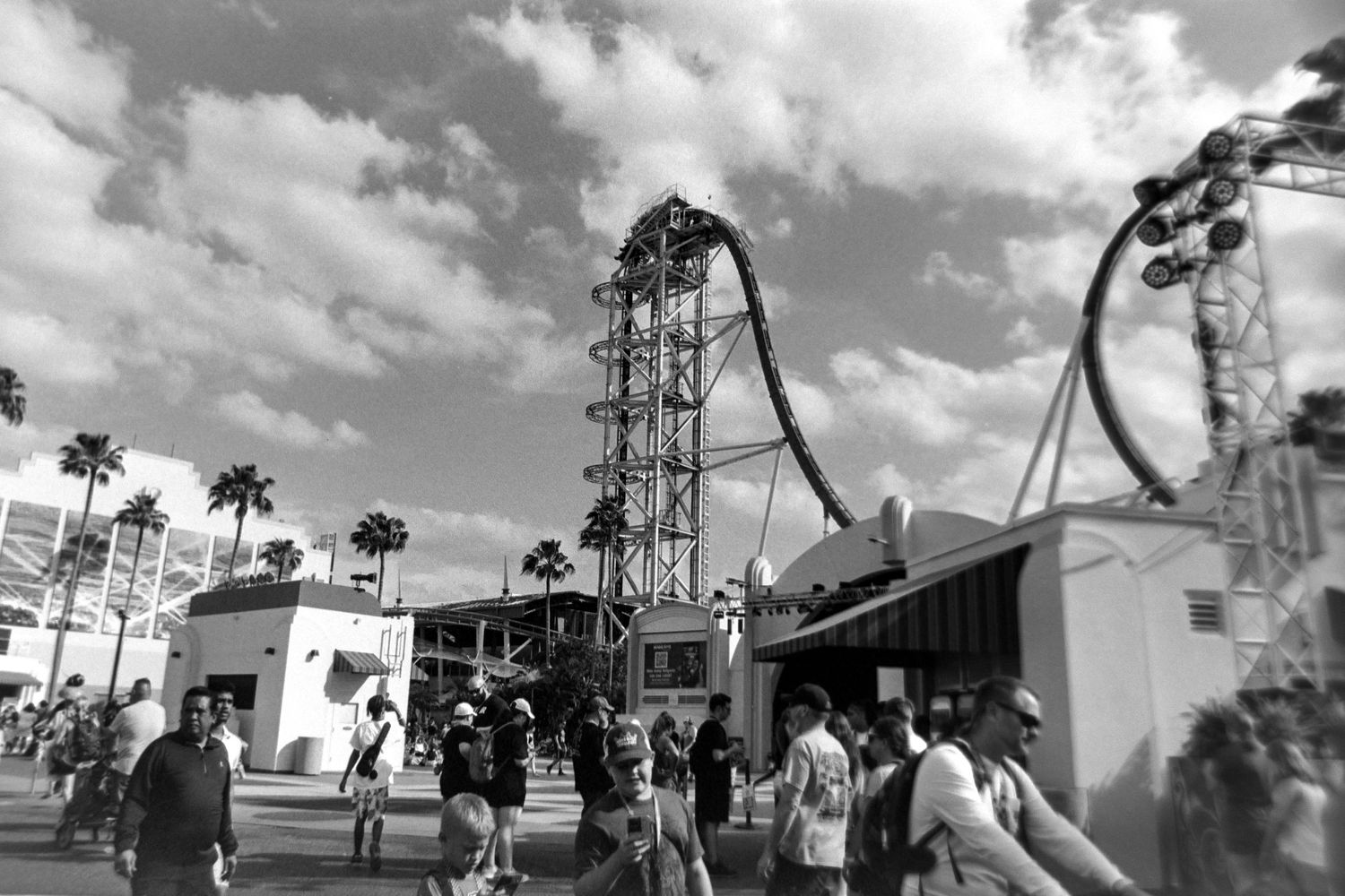 People walking near a towering roller coaster track against cloudy sky at an amusement park in black and white.