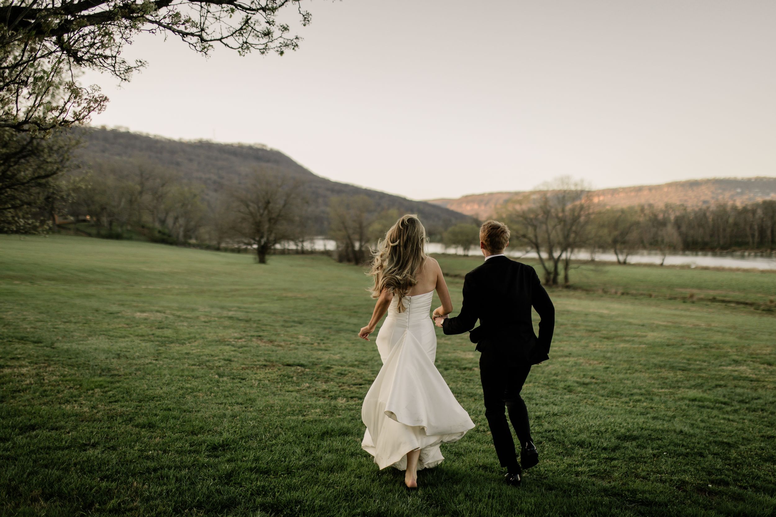 A couple runs through a scenic grassy field with mountains and trees in the background on their wedding day.