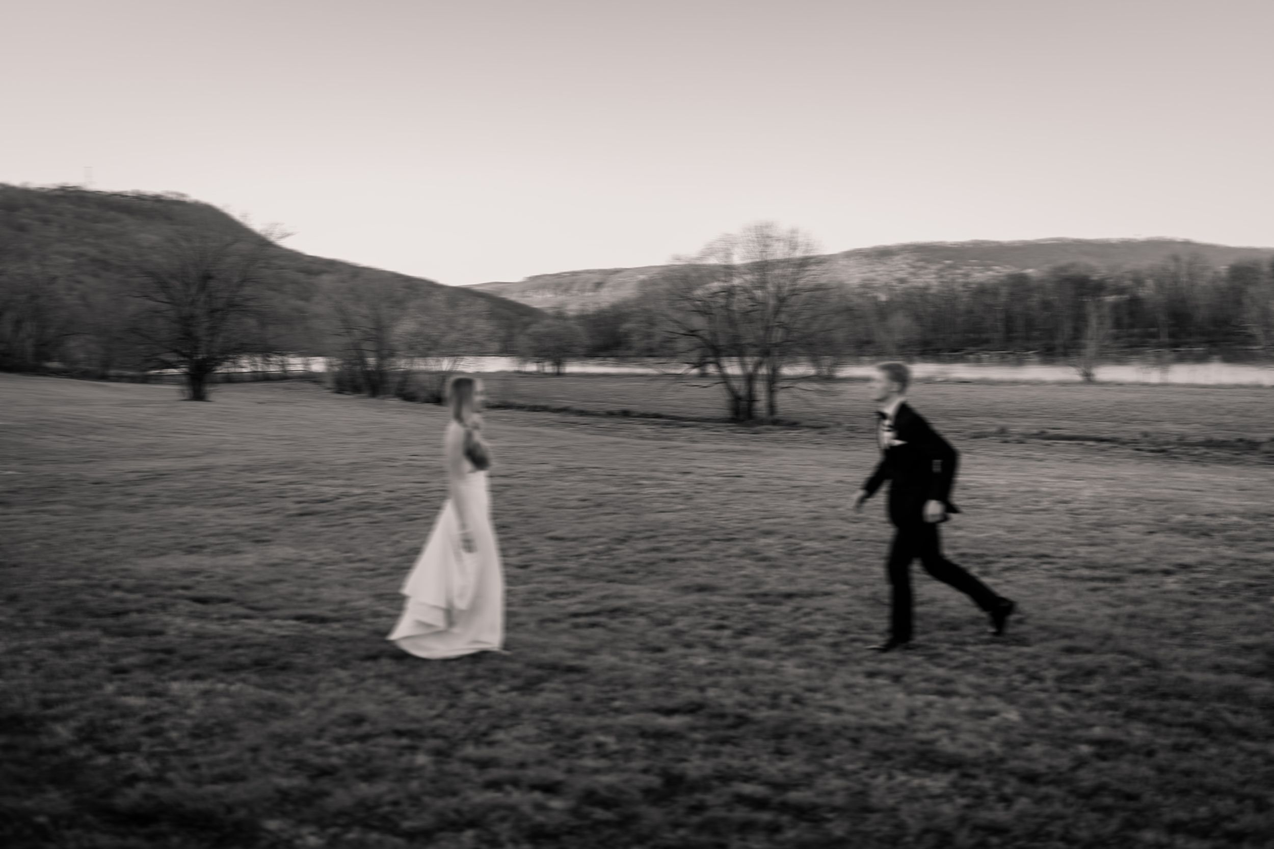 Black and white photo of a playful chase scene at a wedding across a scenic field with mountains in the background.