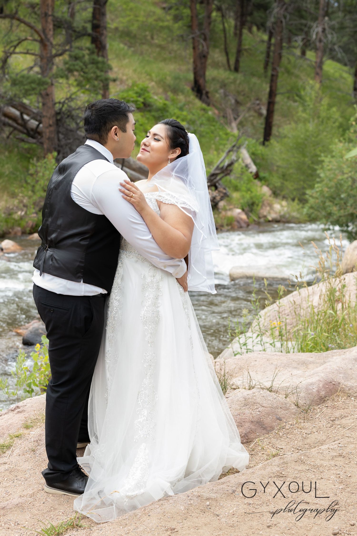 Bride and groom share an intimate moment by a mountain stream in their wedding attire.