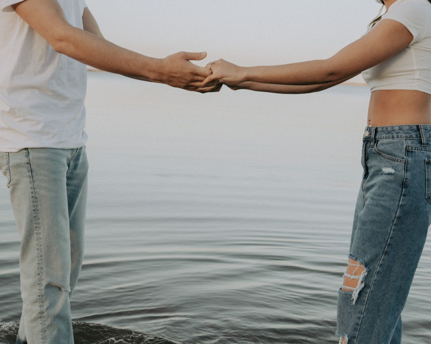 Two people in jeans and white shirts holding hands while standing near calm water.