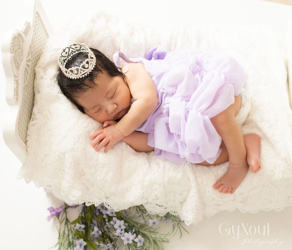 Newborn in a lavender tutu dress sleeping peacefully on white lace with purple flowers.