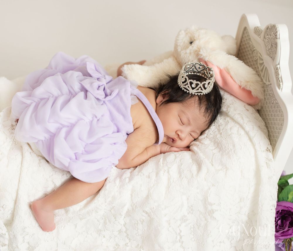 Sleeping infant wearing lavender tulle dress and silver crown on white bedding.