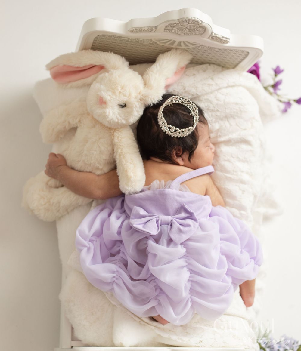 Sleeping baby in purple dress with white bunny plush on lace background.