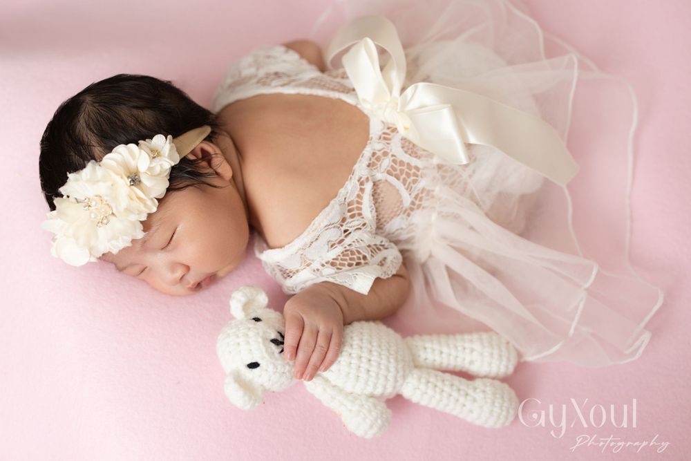 Sleeping newborn in white lace outfit with floral headband holding knitted teddy bear.
