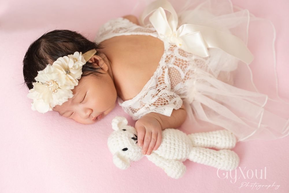 Peaceful sleeping baby in delicate white lace dress clutching small knitted bear.
