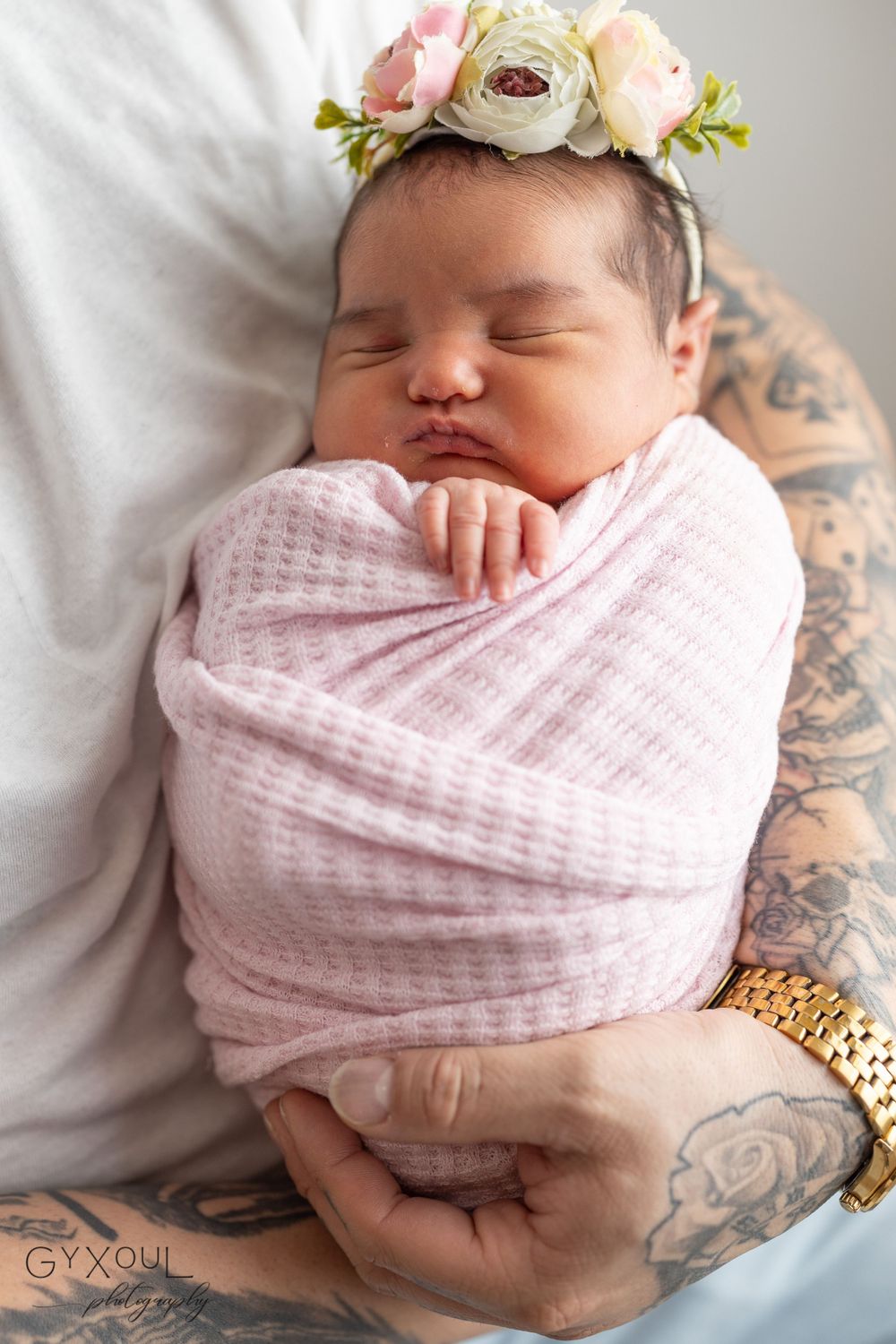Sleeping newborn wrapped in pink swaddle wearing delicate flower crown with tattooed hands visible.