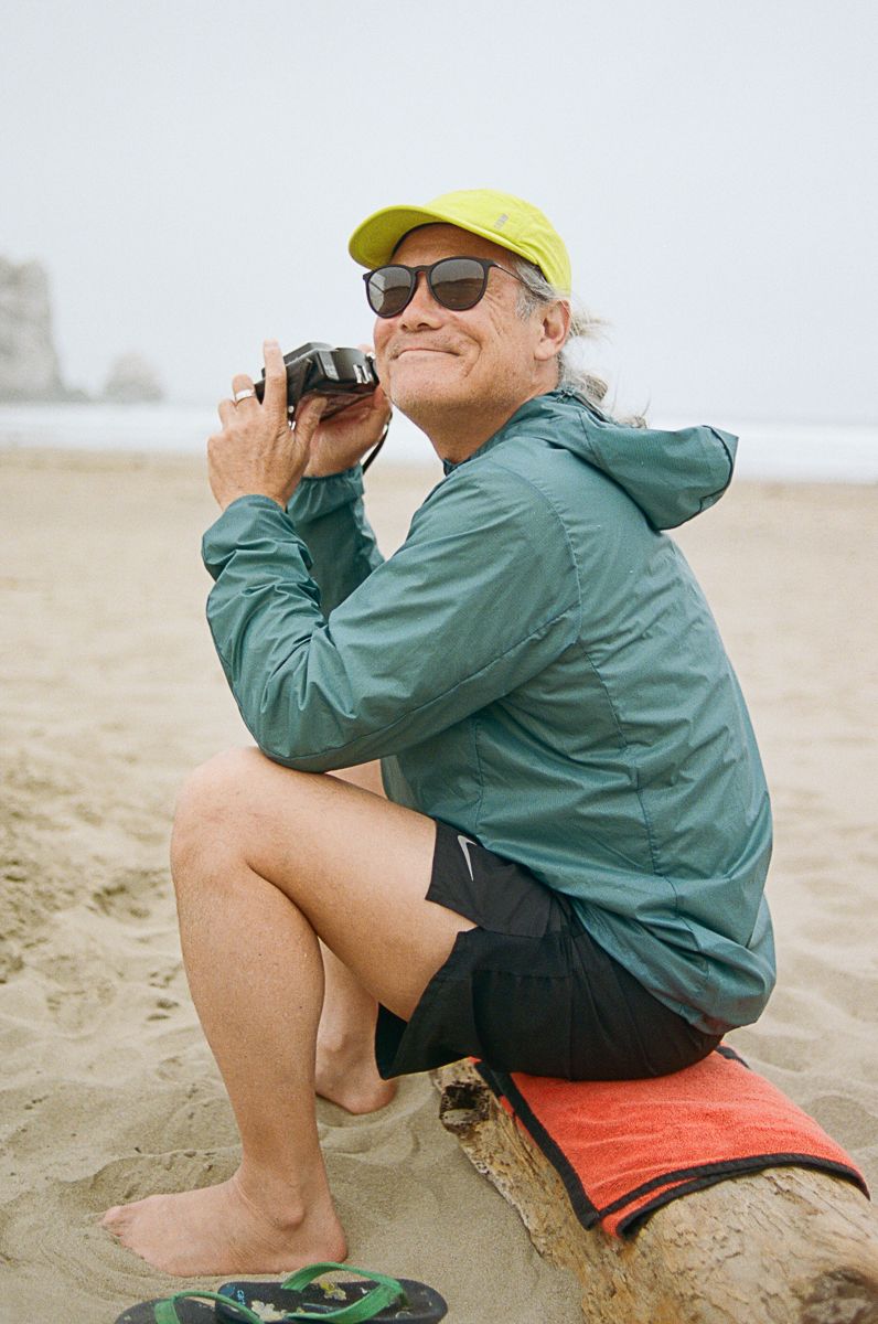 Person in green jacket and yellow cap sitting on beach sand using binoculars.
