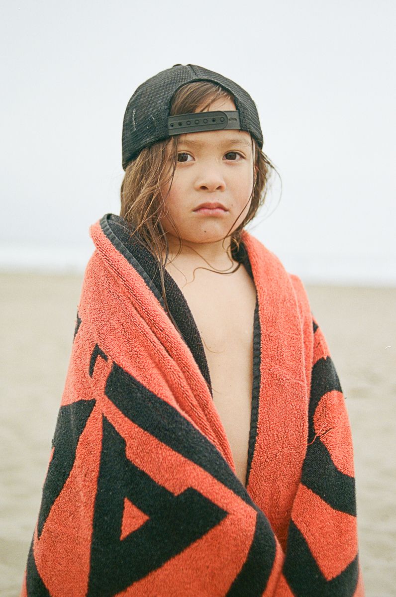 Child wearing an orange and black towel and baseball cap on a misty beach.