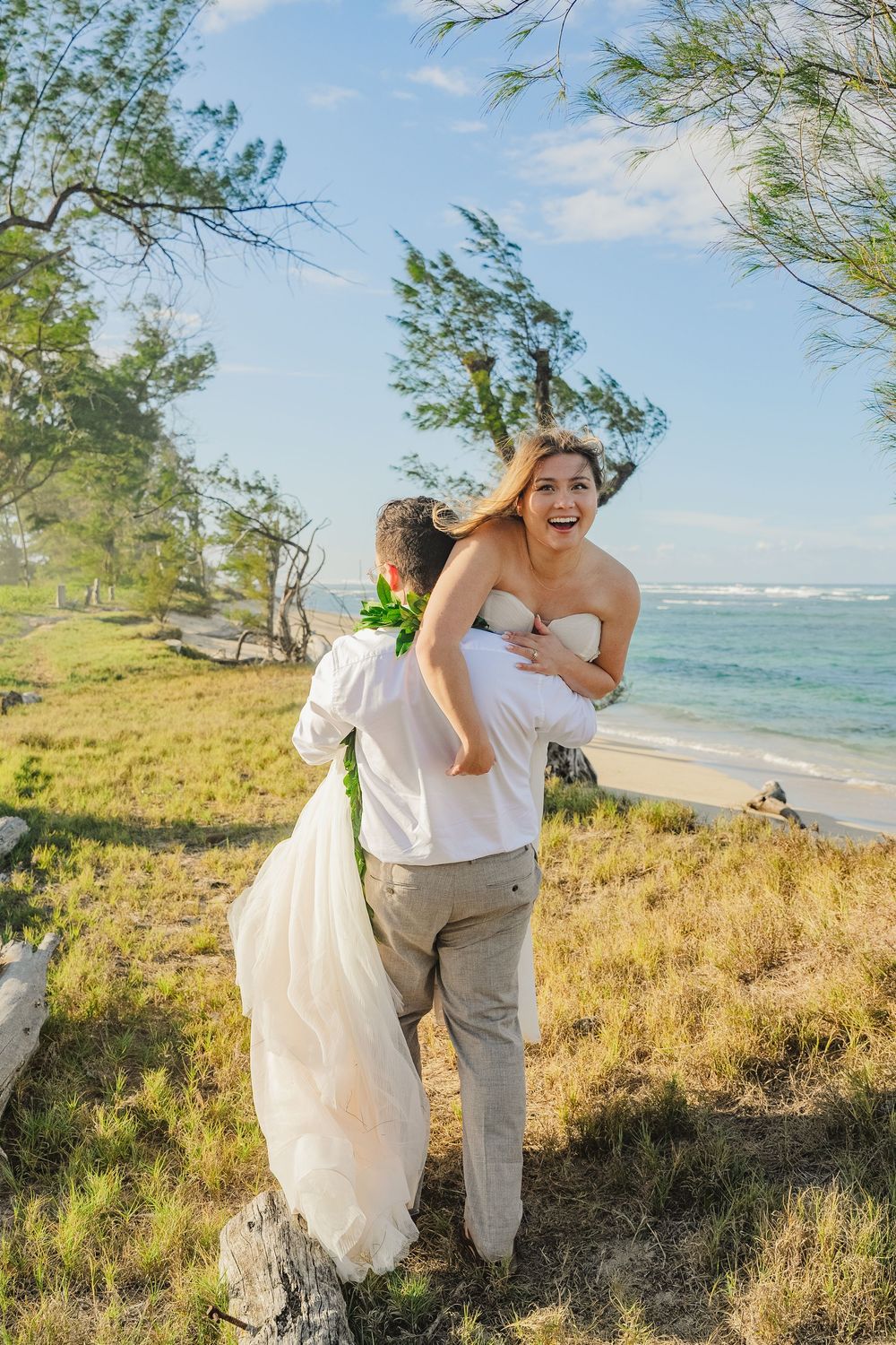 A couple in wedding attire embraces and laughs joyfully on a scenic beach with ocean waves and green foliage in Hawaii.