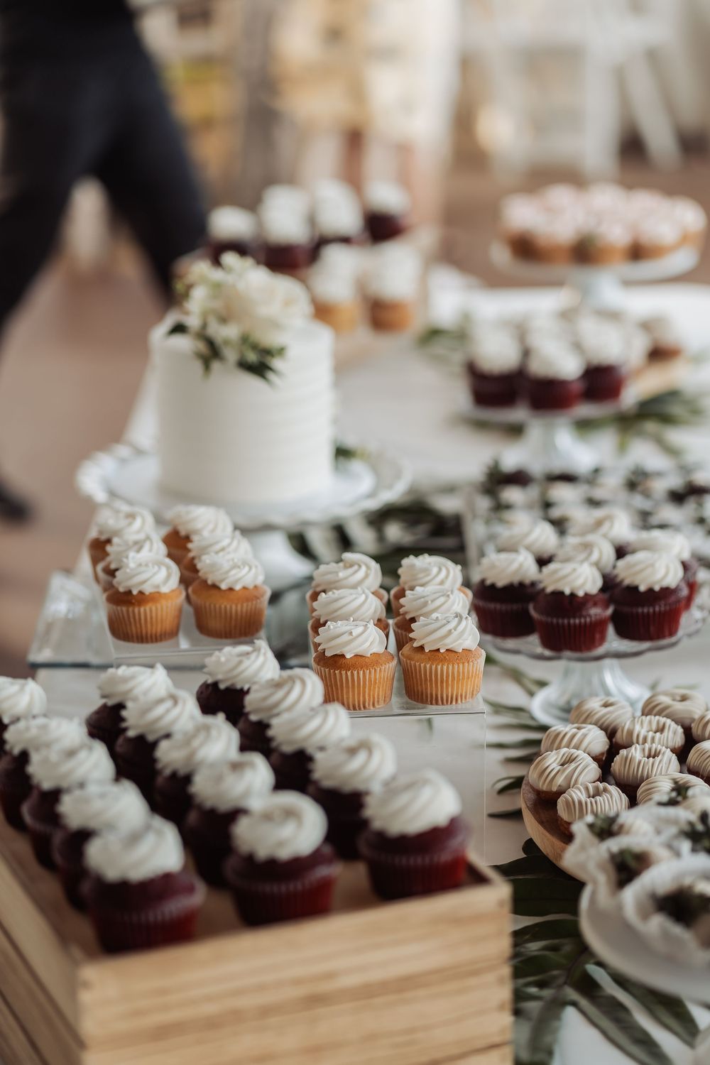 Elegant dessert table display with white frosted cupcakes, chocolate cupcakes and small white cake decorated with greenery.